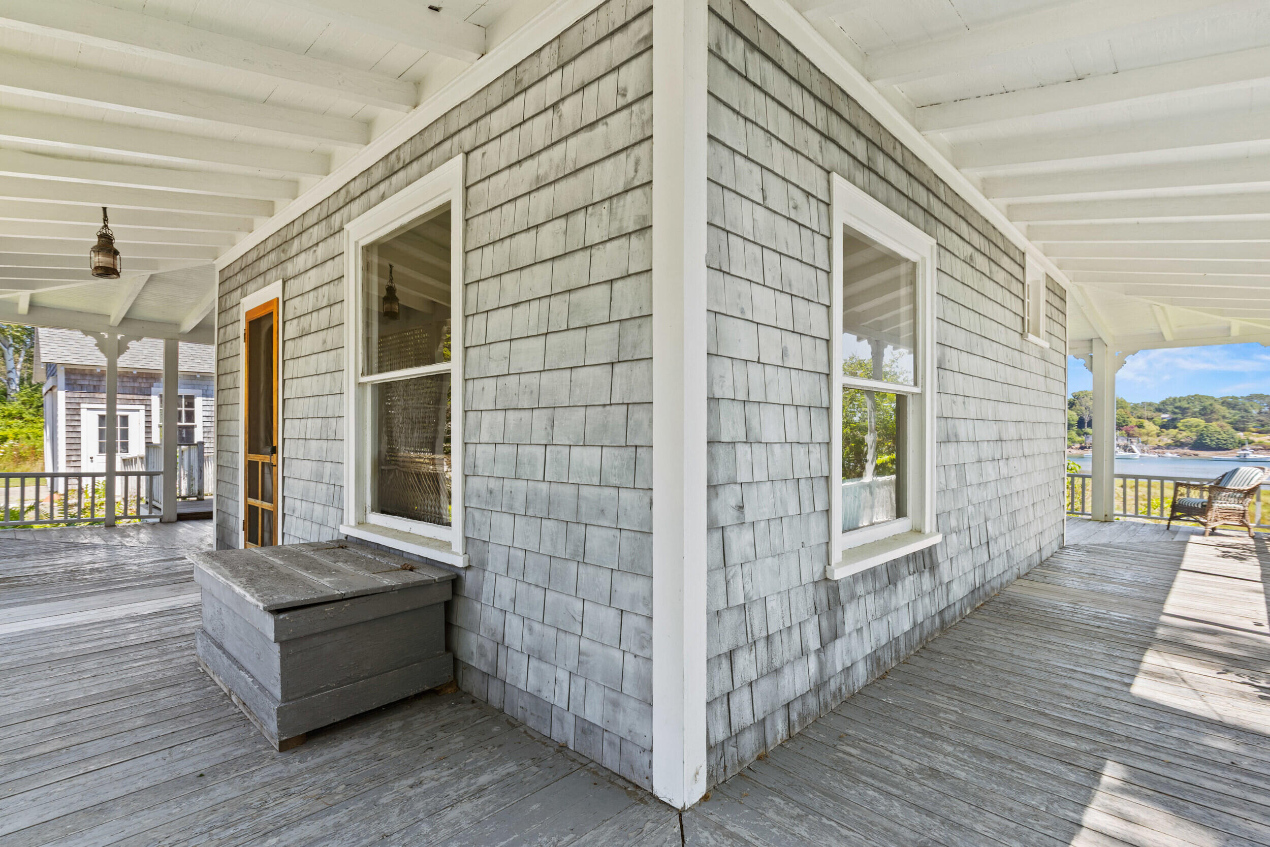 Covered wraparound porch with gray shingles, a wicker chair, and a view of the water.