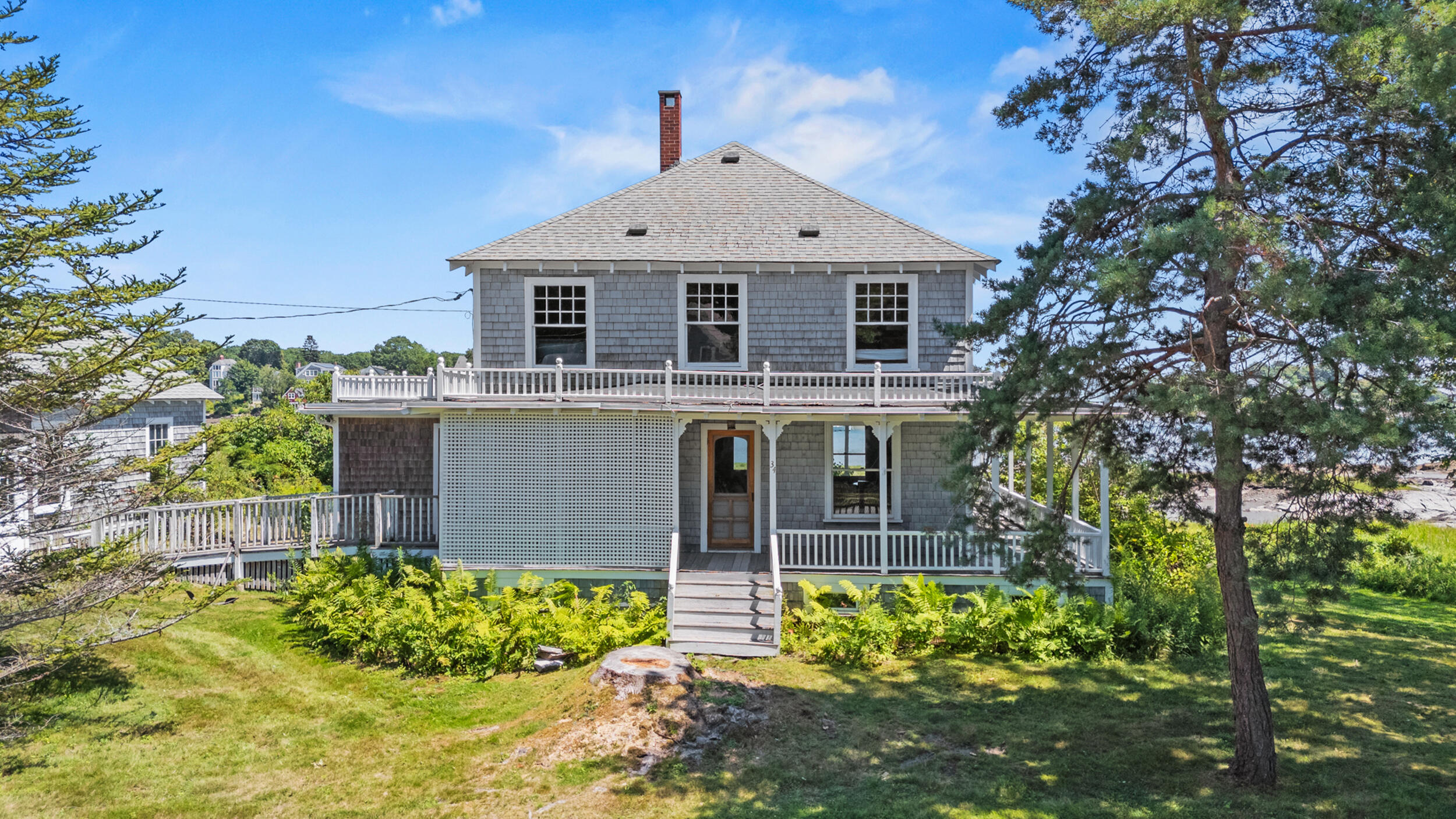 Gray shingle house with a broad porch and front steps, framed by trees and ferns.