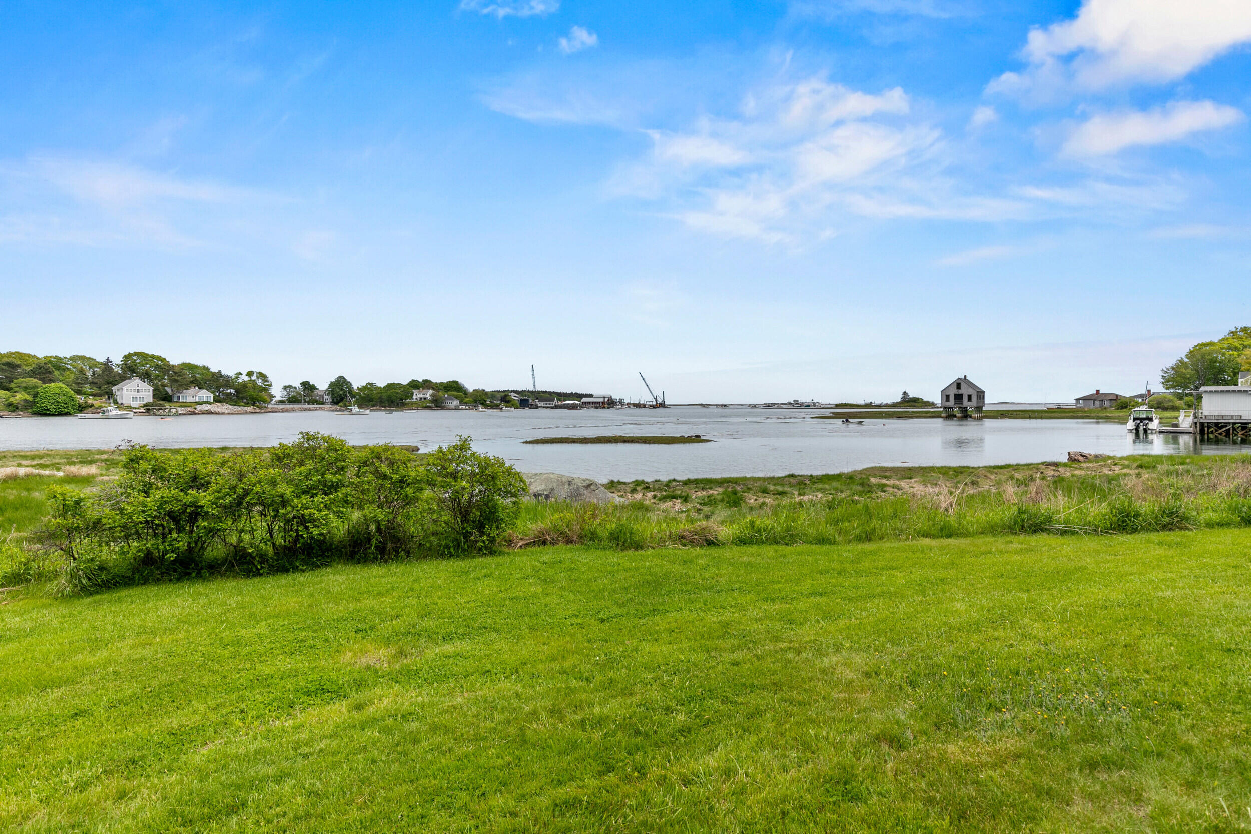 Tidal inlet and shoreline homes beyond a broad green lawn, with boats and a small stilt house.