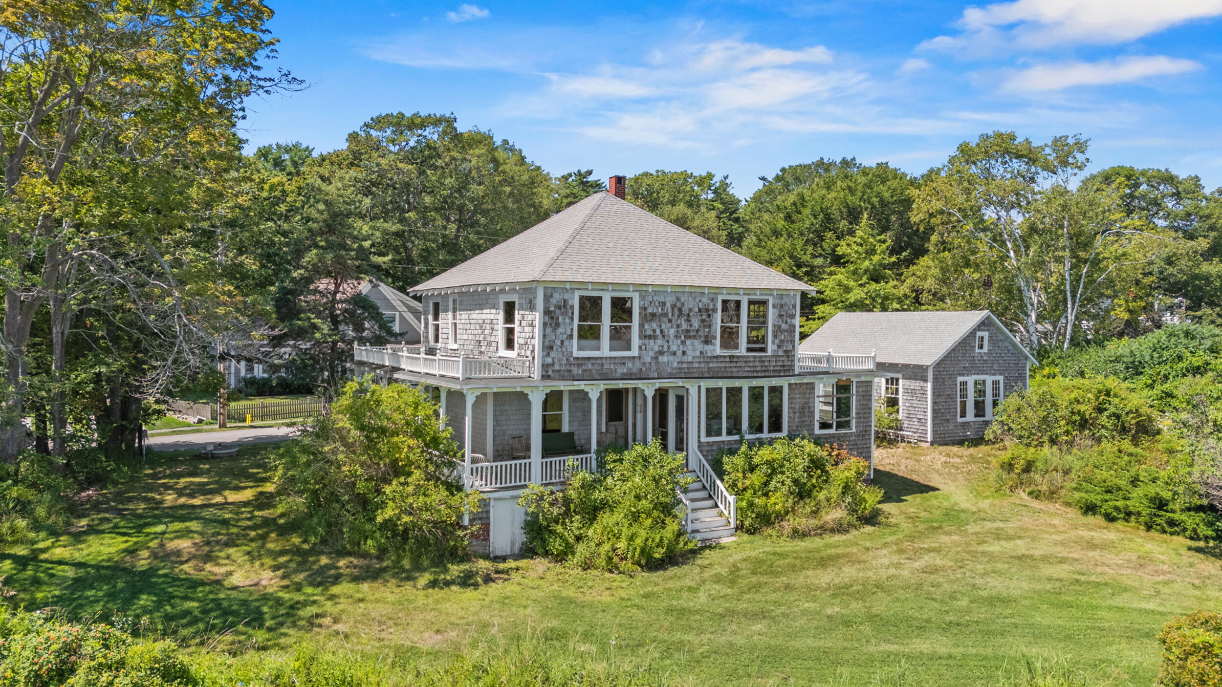 Shingle-sided two-story house with a covered porch and attached wing amid mature trees.