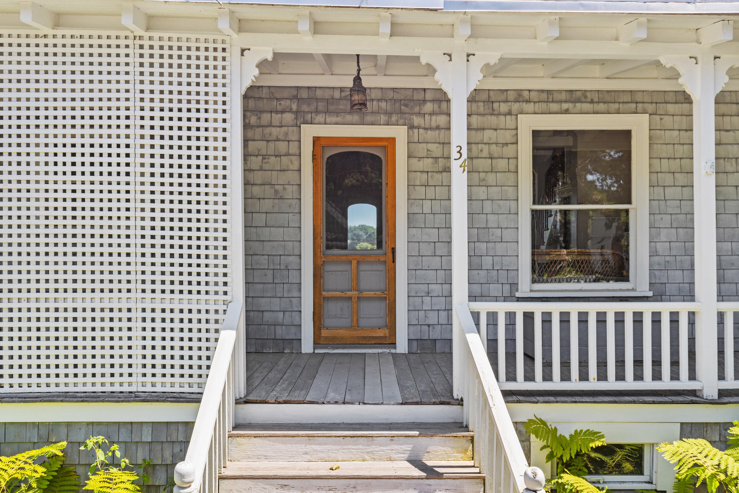 Front porch with gray shingles, a wood-framed screen door, white lattice, and ferns beside the steps.