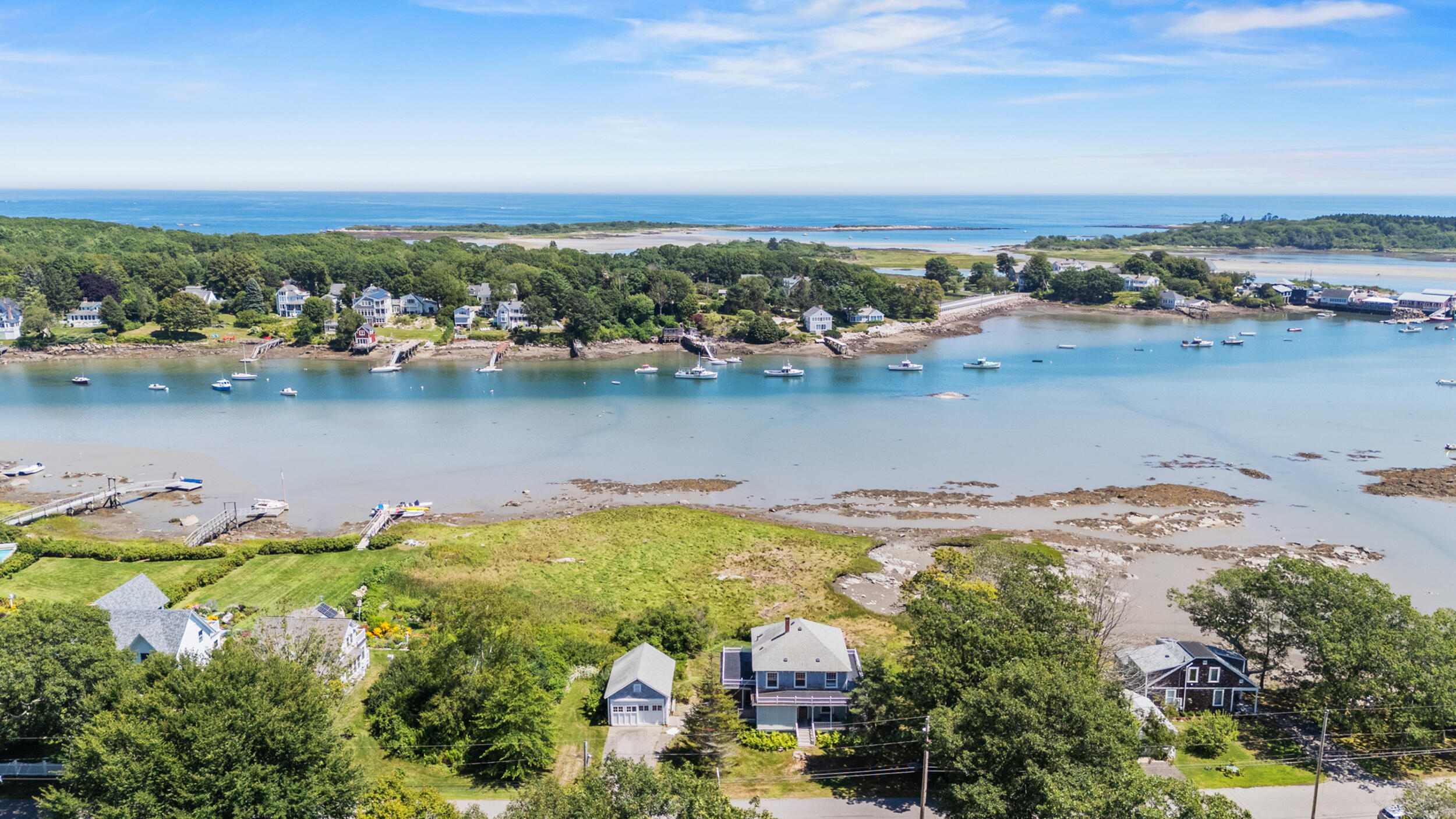 Aerial view of a coastal home beside a boat-filled tidal harbor and the open ocean.