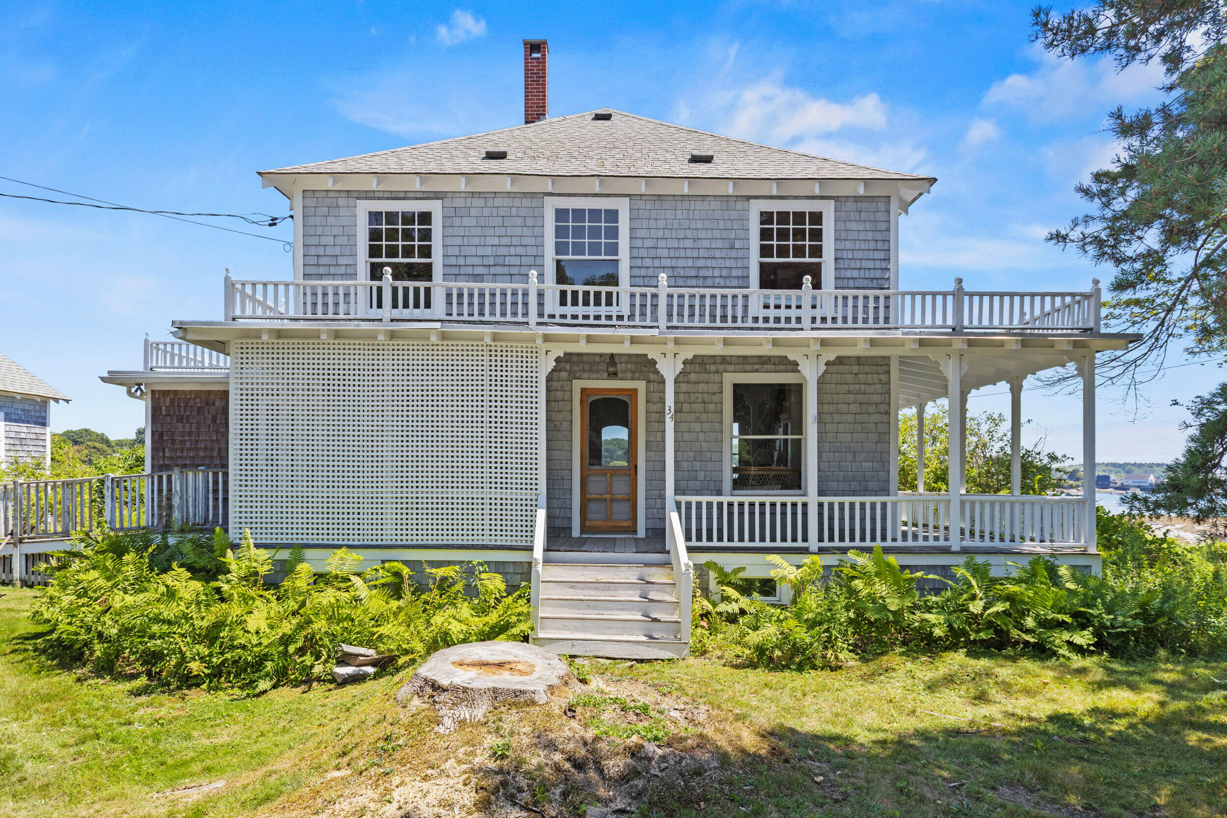 Two-story gray-shingled house with white wraparound porches and water glimpsed through trees.
