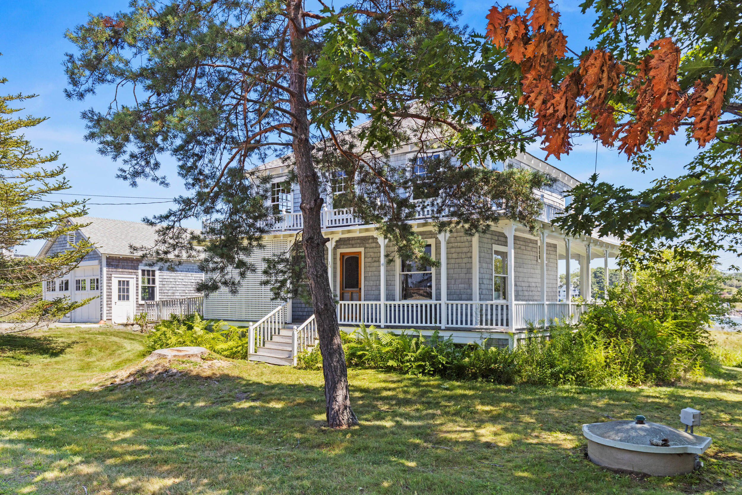 Gray shingle-sided house with a white wraparound porch, framed by mature trees and a green lawn.