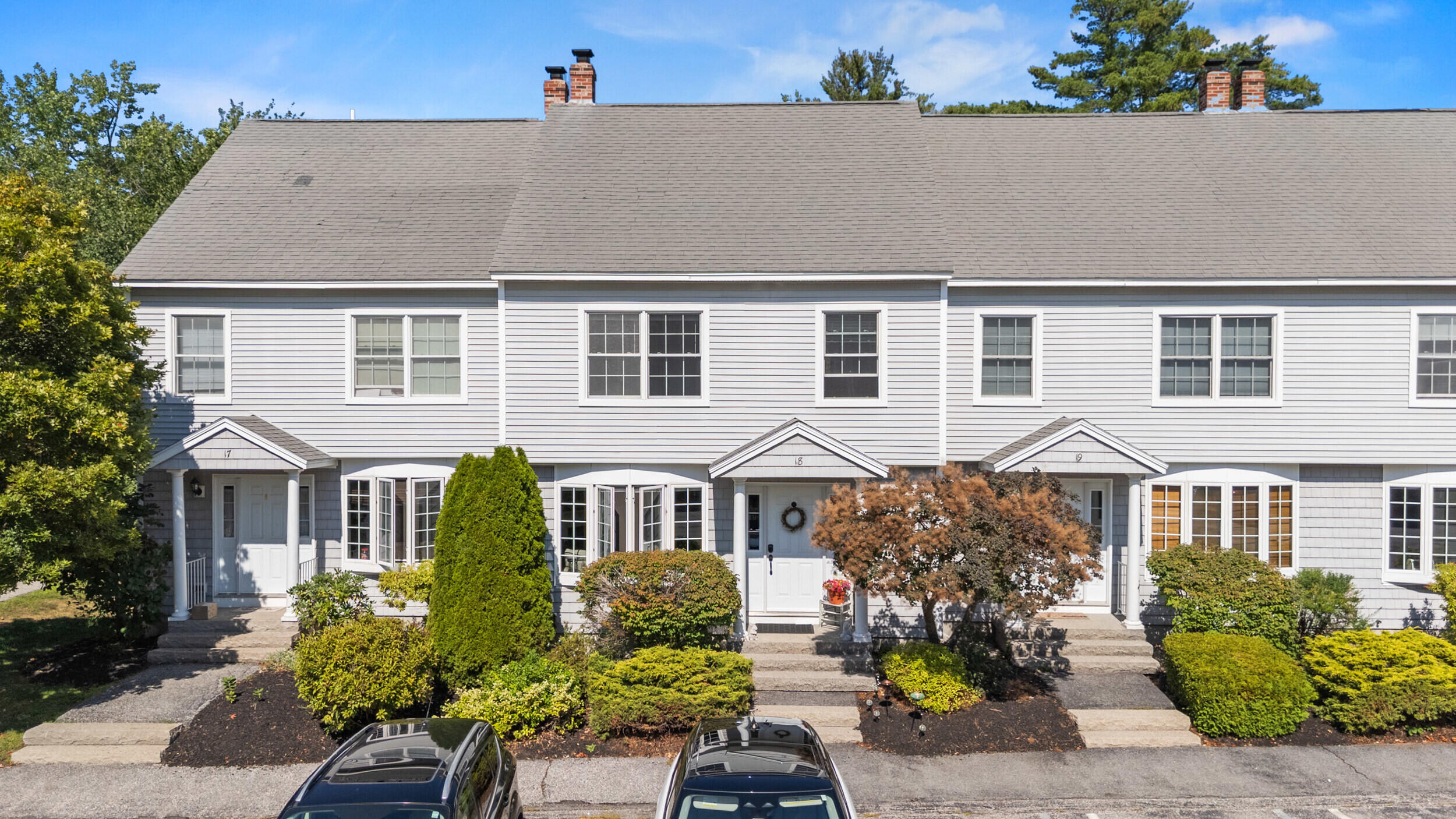 Row of gray-sided homes with gabled entry porches, landscaped shrubs, and cars parked in front.