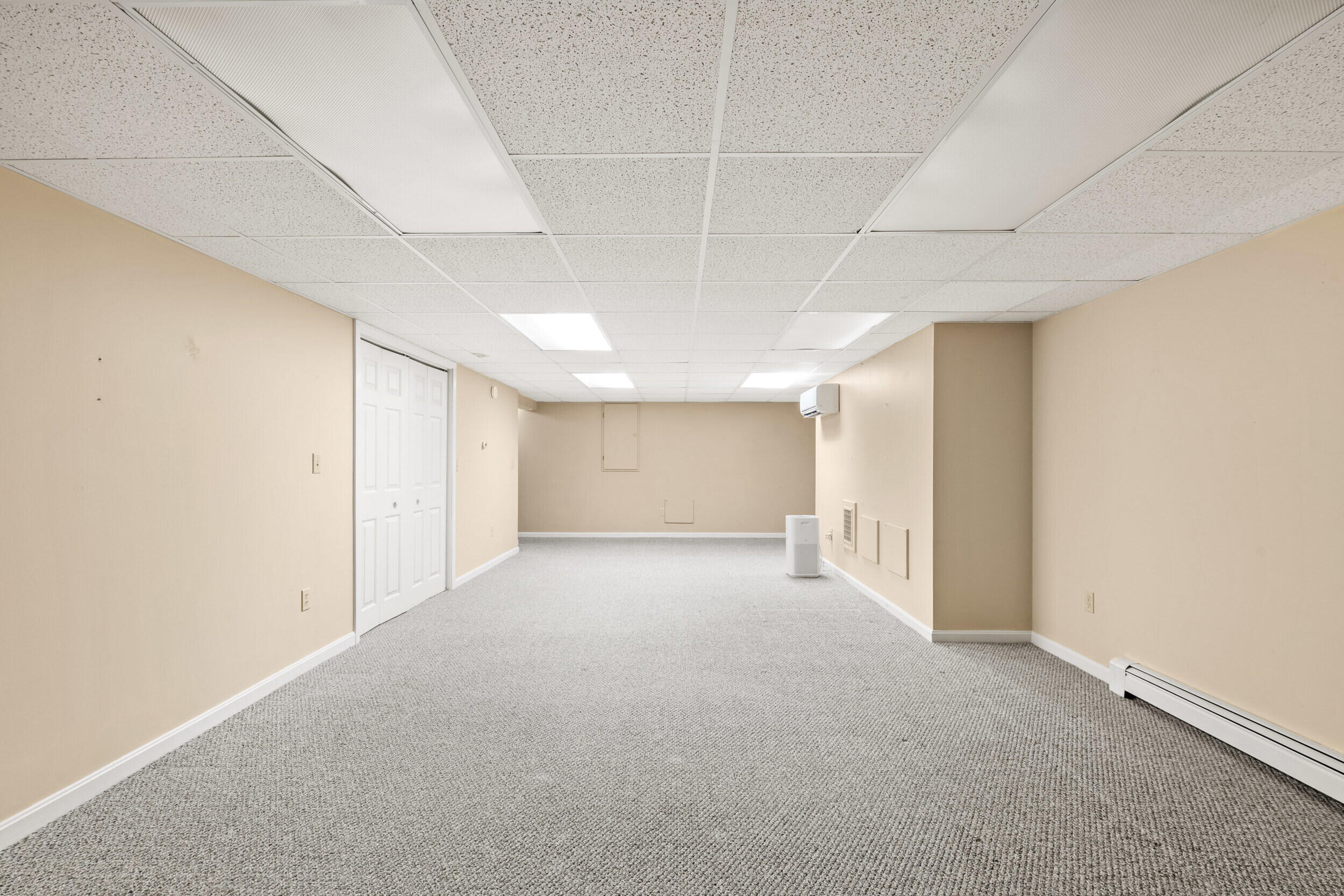 Empty carpeted lower-level room with beige walls, white closet doors, and a suspended ceiling.