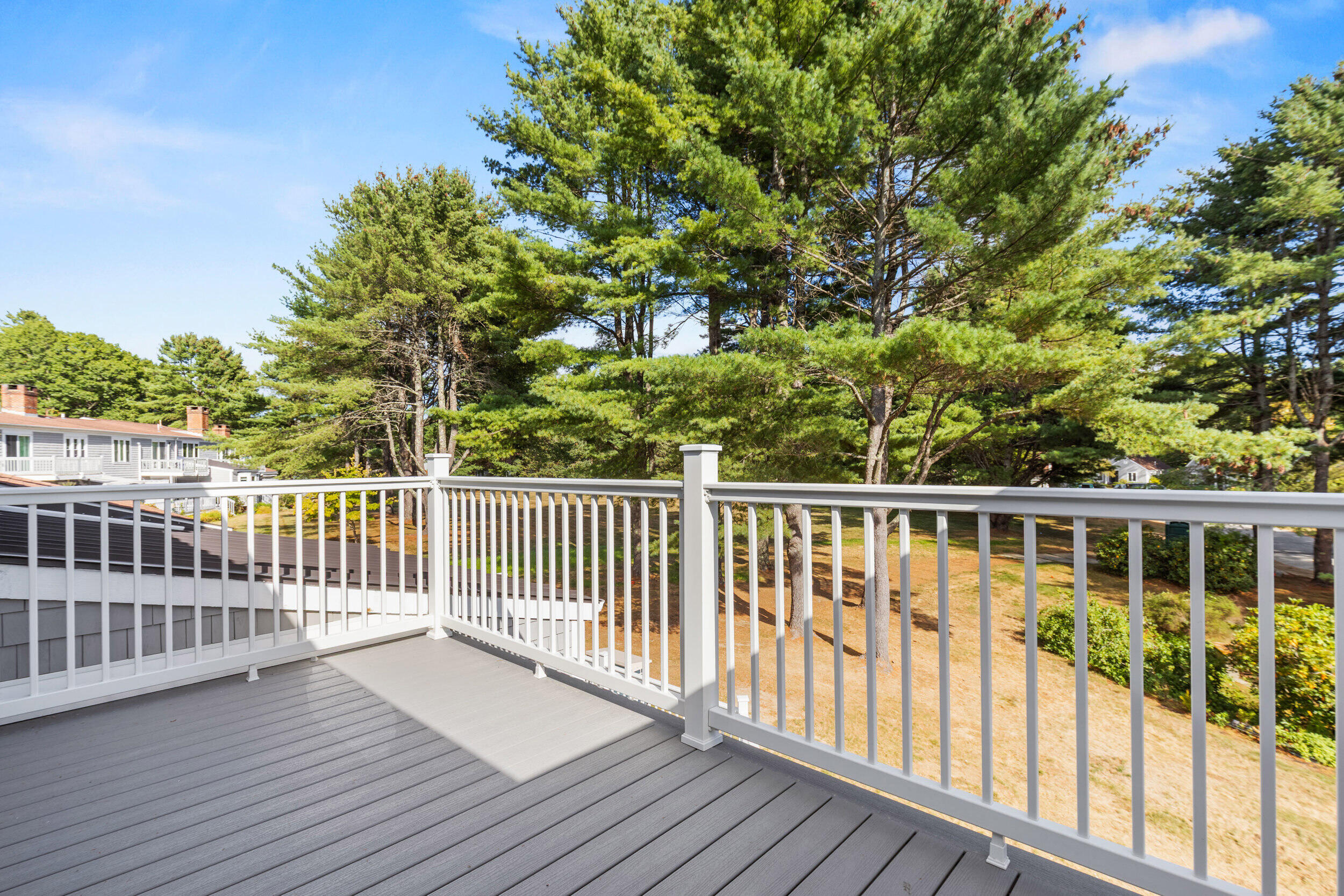 Gray deck with white railings overlooking a sunny yard and tall pine trees.
