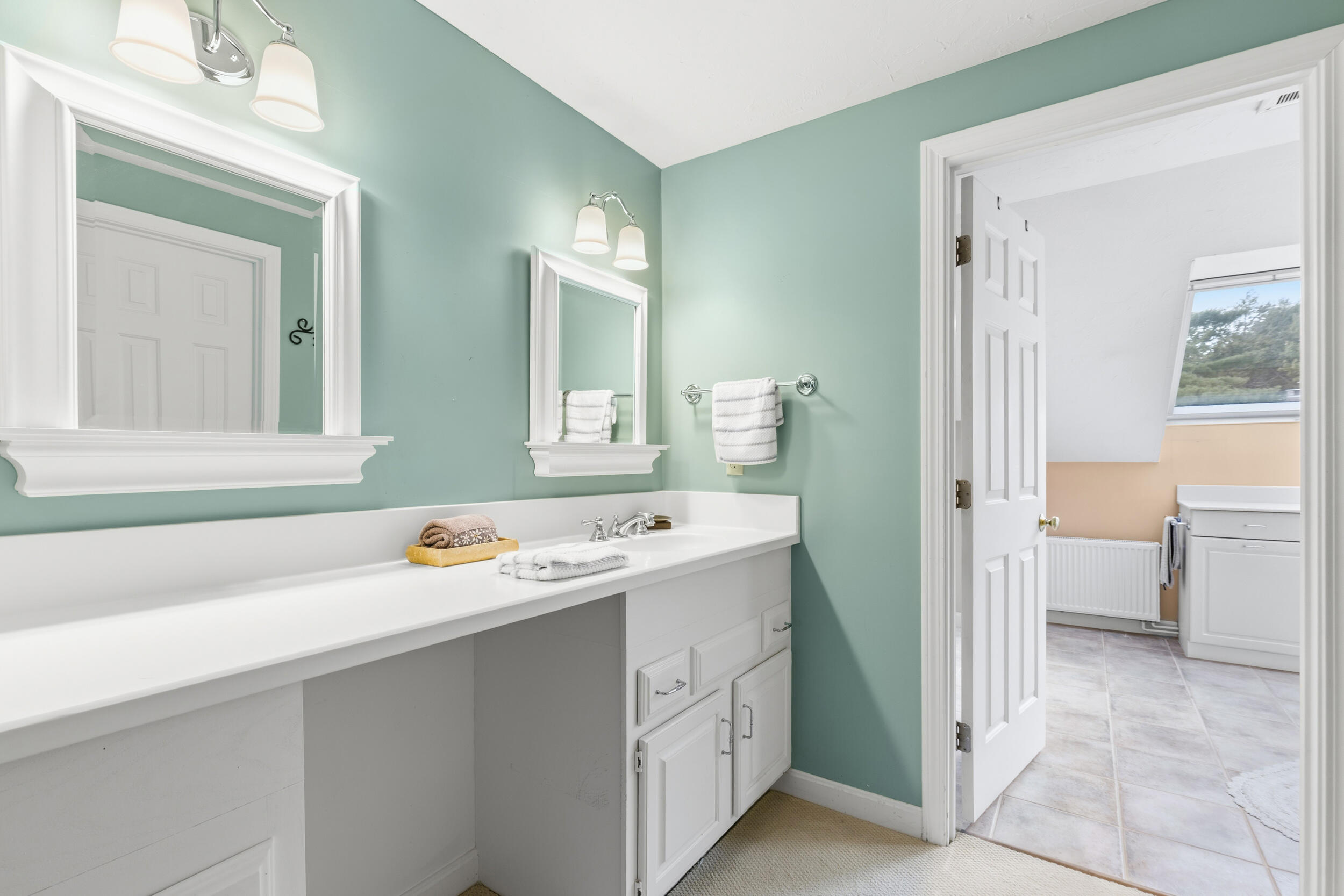 Bathroom with a long white vanity, two mirrors, pale green walls, and an open doorway to a tiled room.