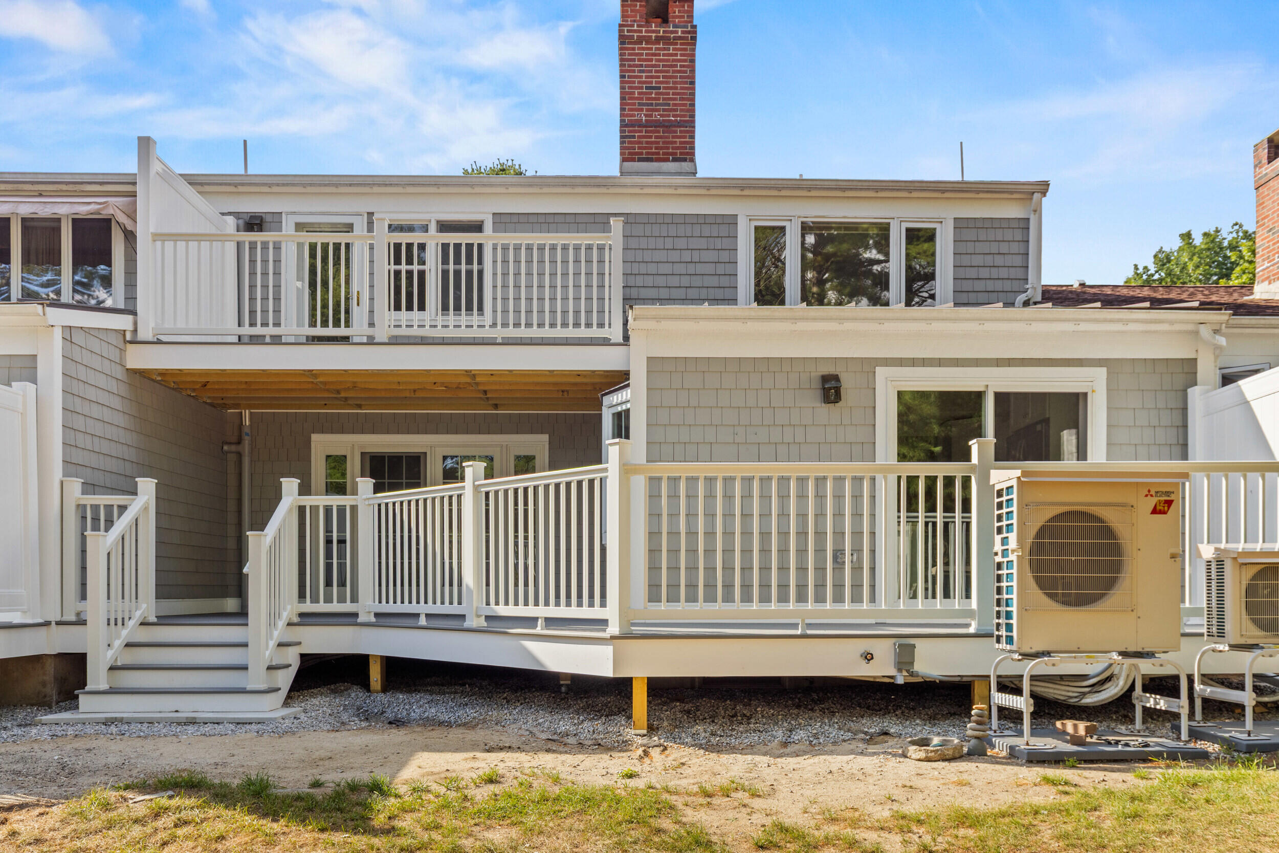 Rear of a gray-shingled house with tiered white decks, stairs, and outdoor heat pump units.