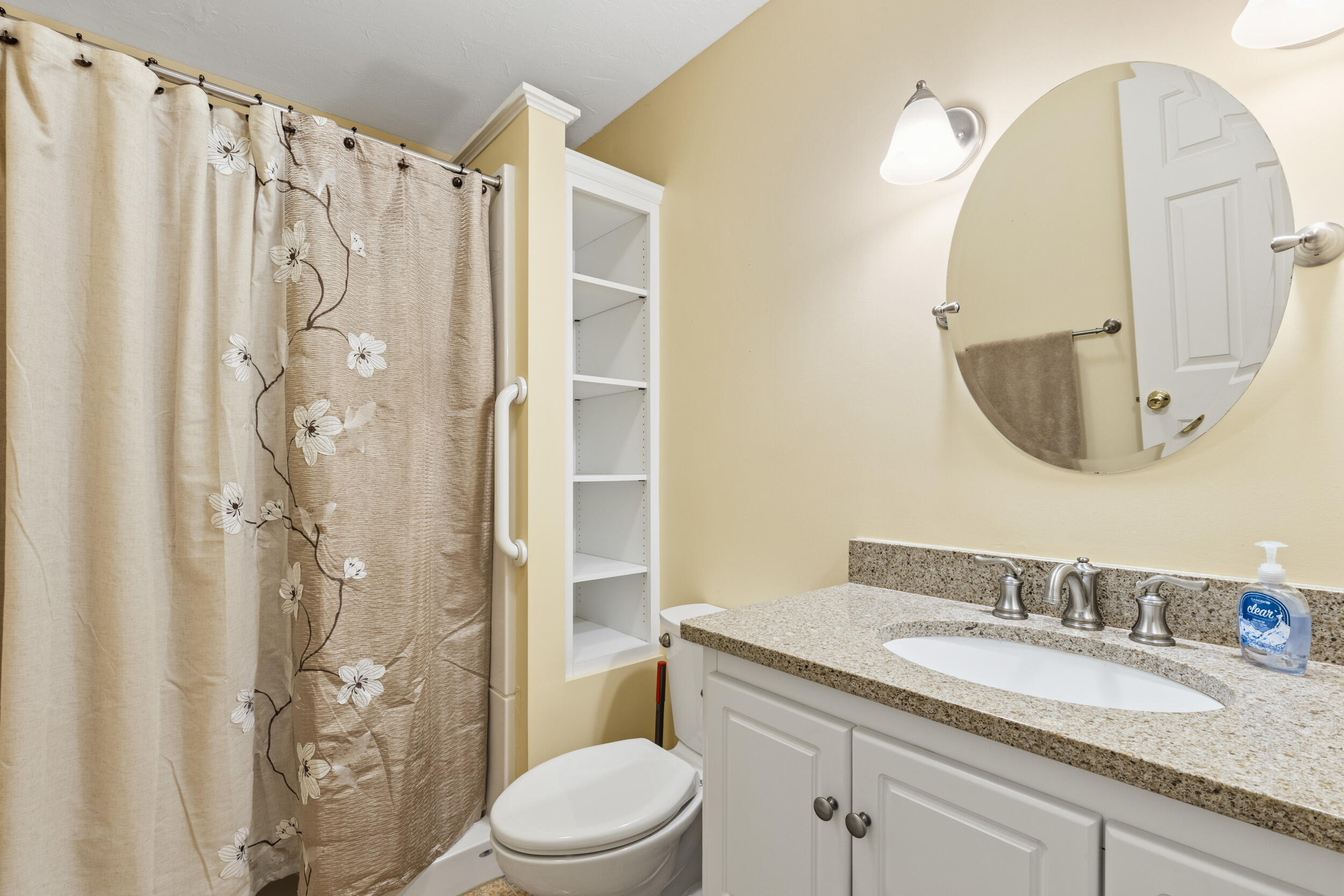 Bathroom with a floral shower curtain, granite vanity, oval mirror, and built-in shelving.