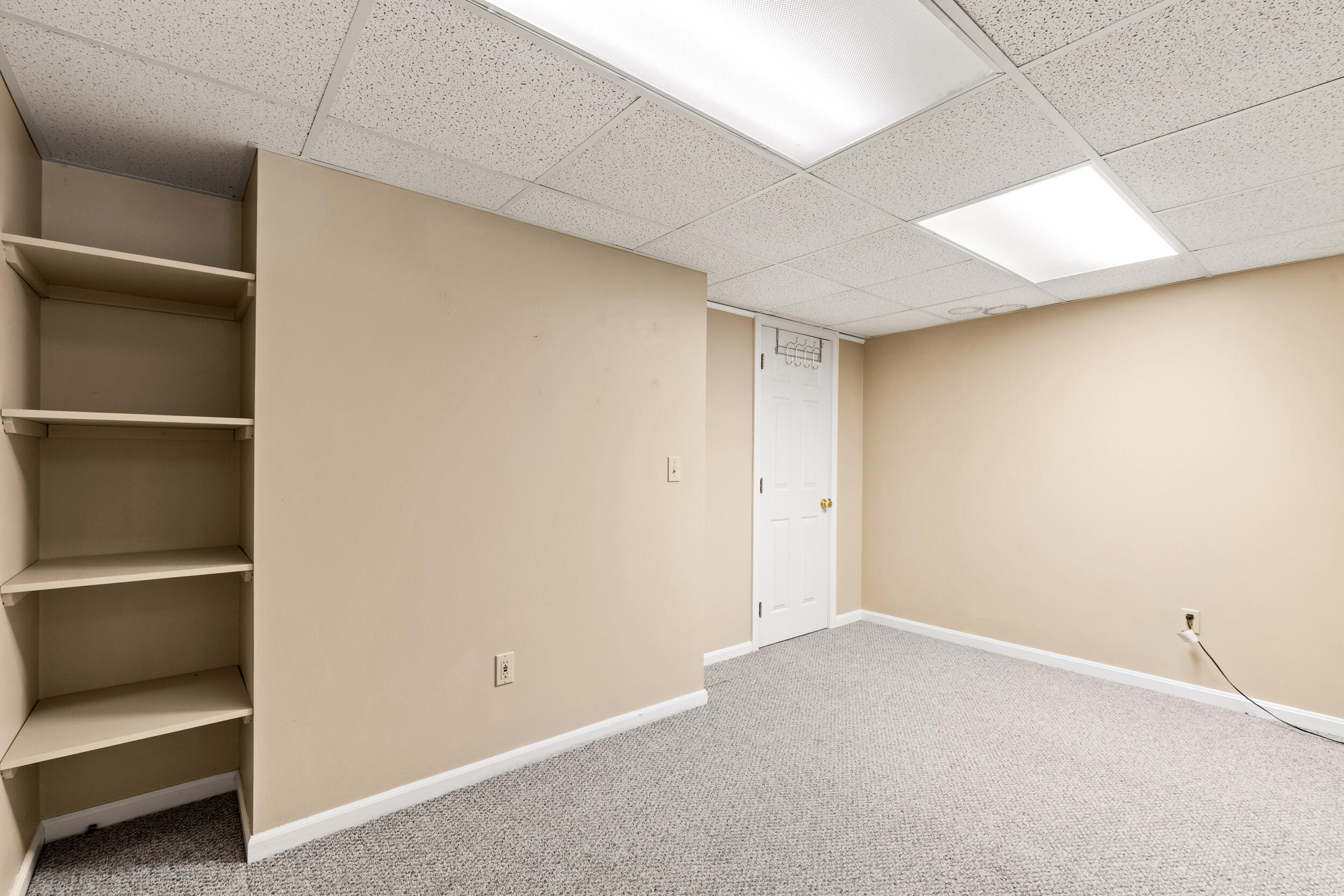 Empty carpeted room with built-in shelving, beige walls, and a white door.