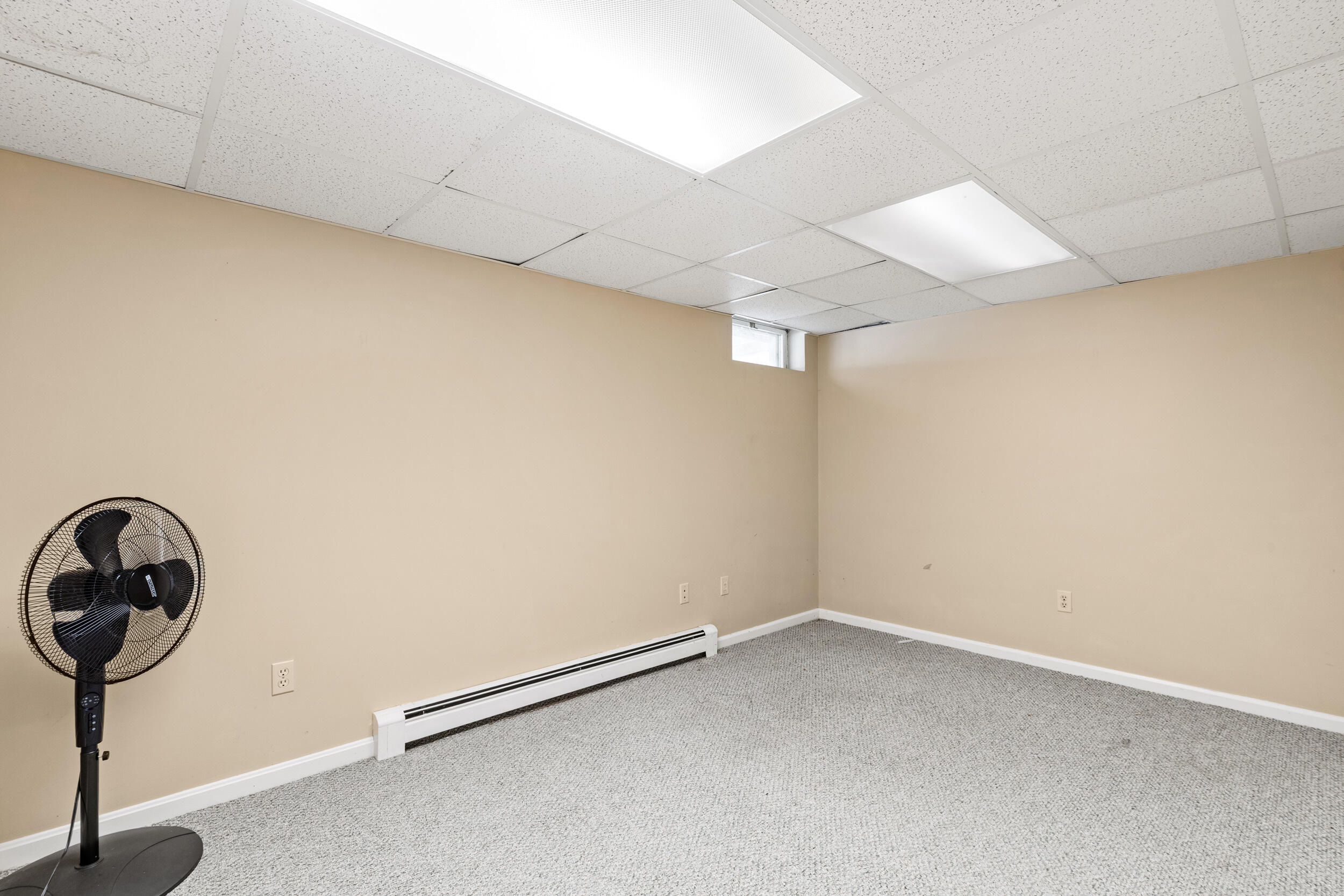 Empty carpeted room with beige walls, fluorescent ceiling lights, and a standing fan.