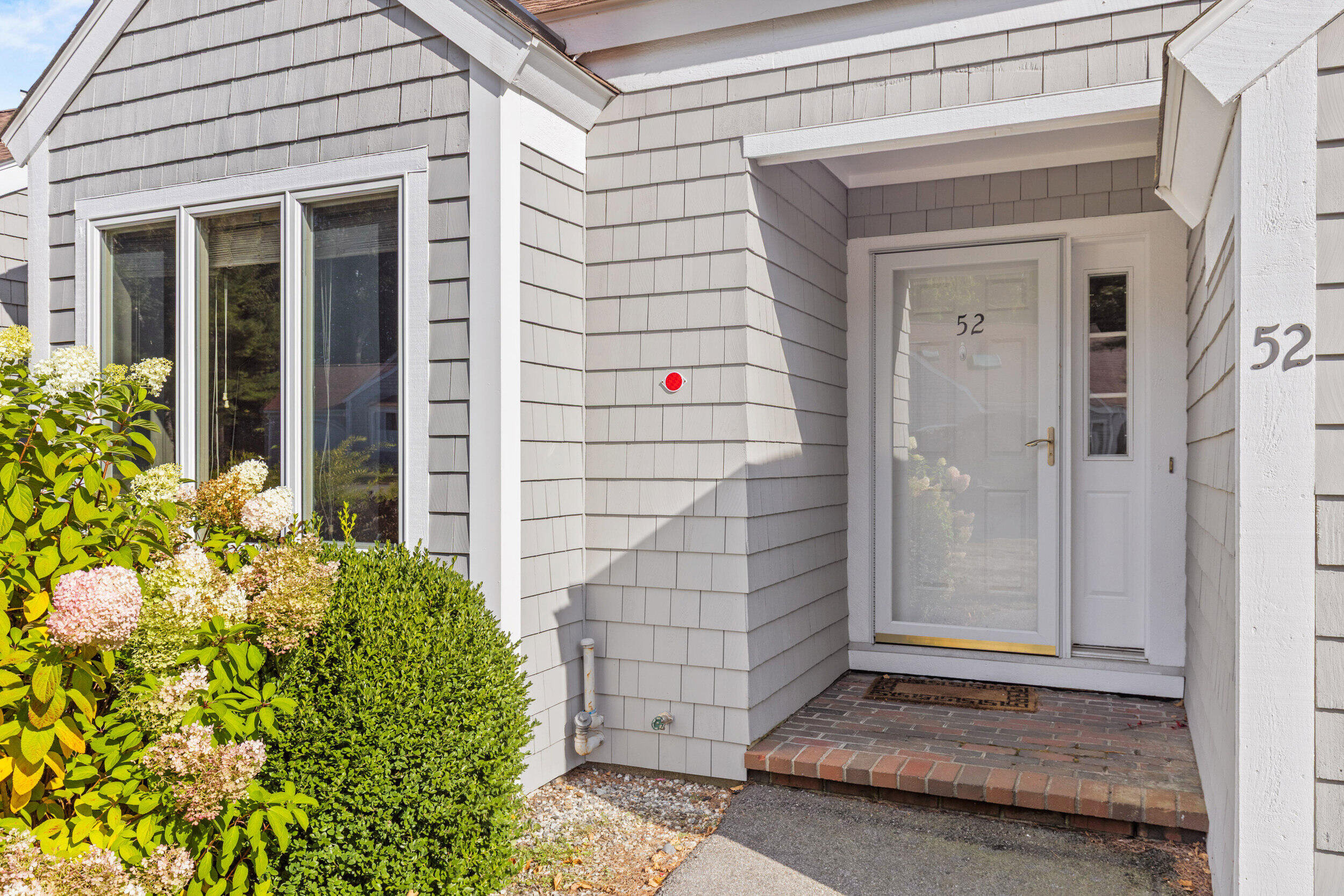 Gray shingled home entrance with a white storm door, brick stoop, flowering shrubs, and the number 52.