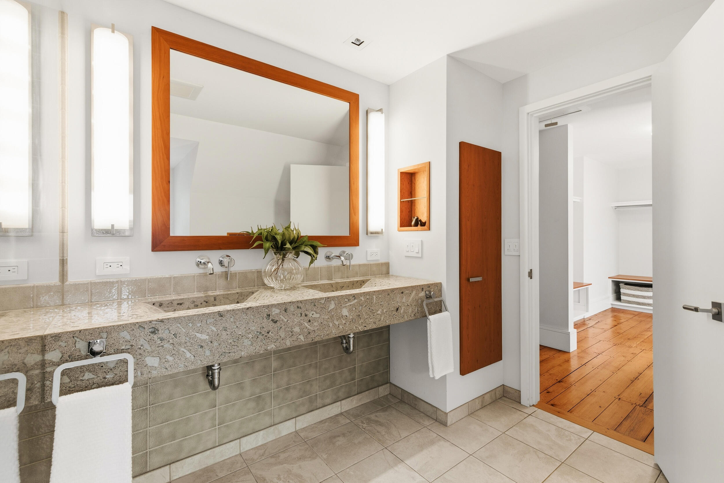 Bathroom with a stone double vanity, wood-framed mirror, and doorway to a room with wood floors.