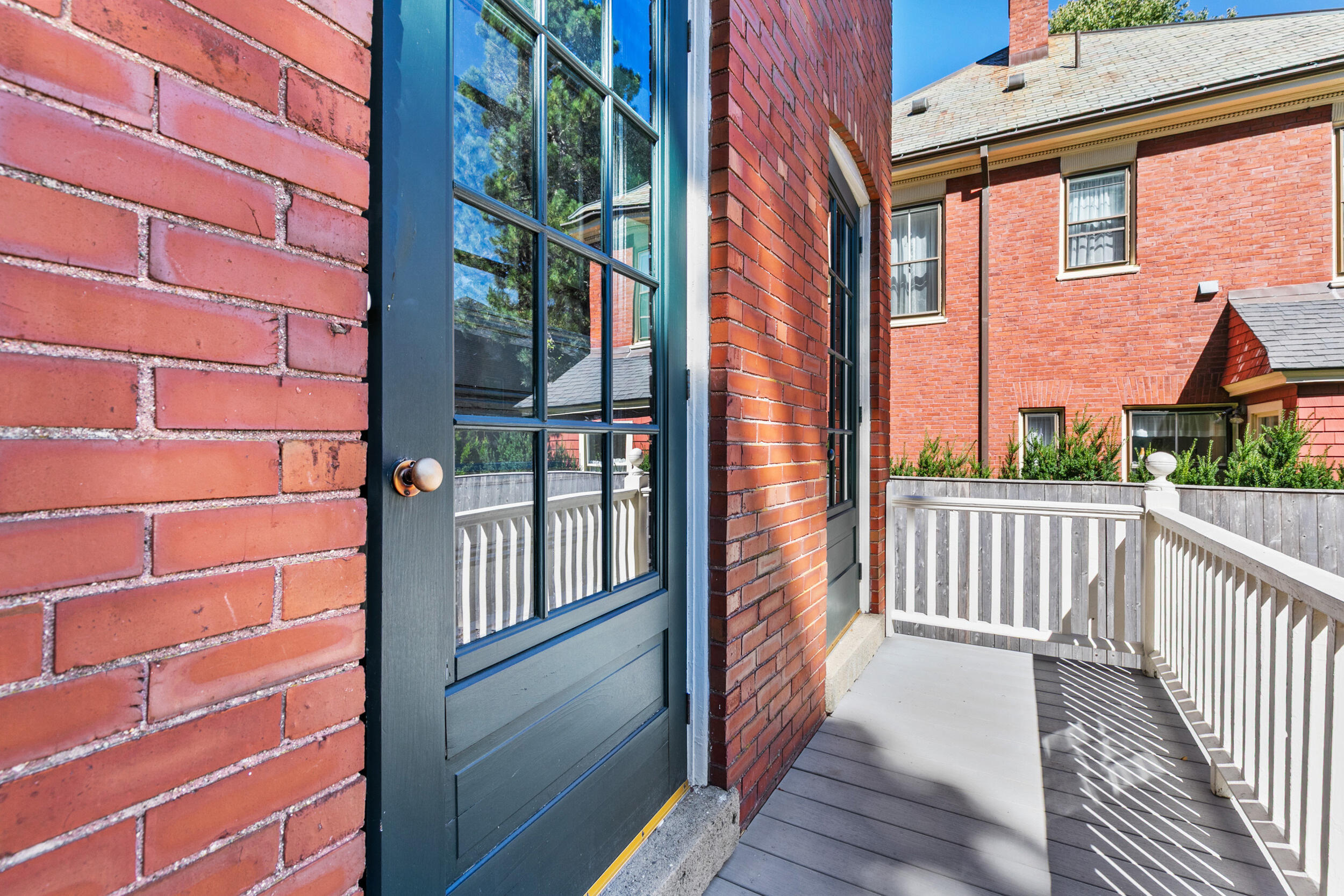Blue glass-paneled entrance door beside a sunny deck with white railing and brick buildings.