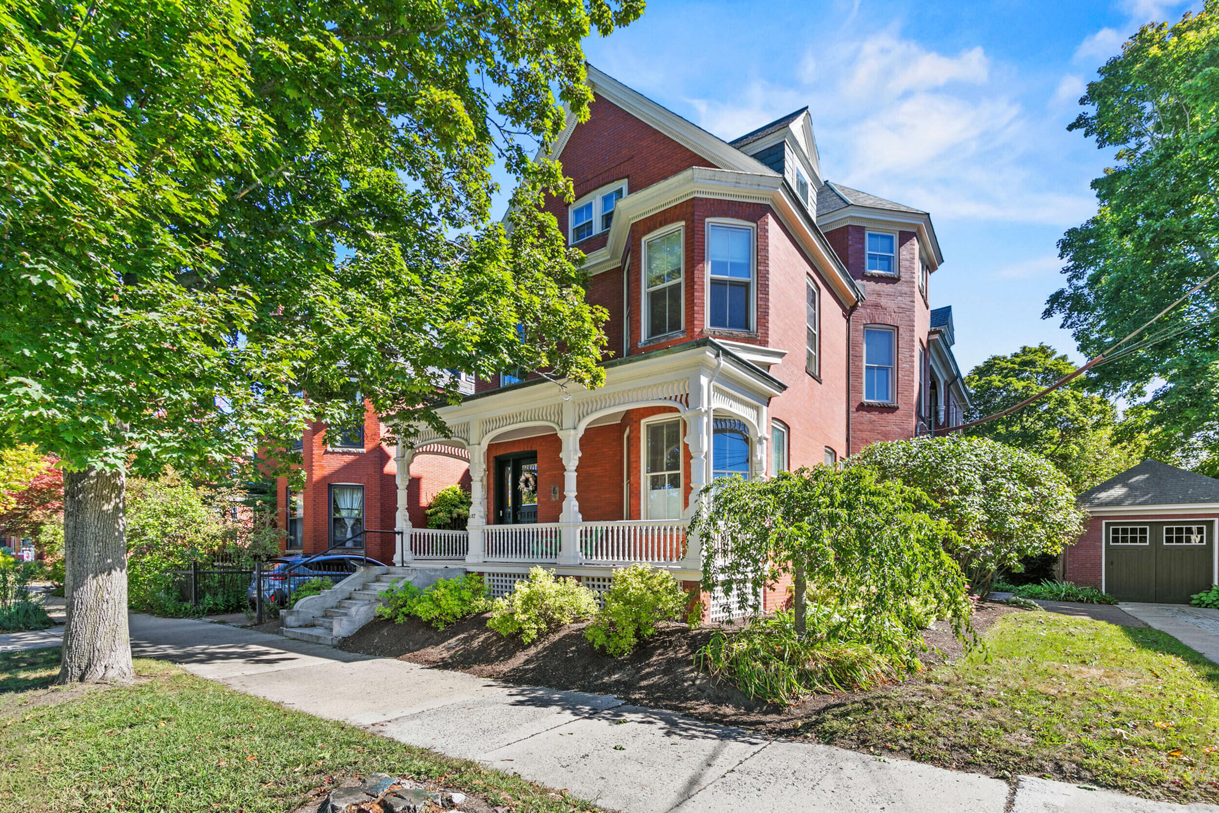 Red brick Victorian home with an ornate white porch, mature trees, and landscaped front garden.