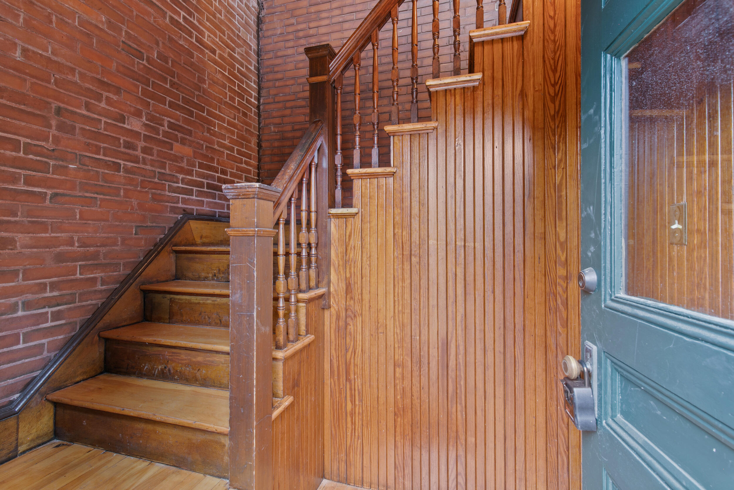 Wooden staircase beside exposed brick walls and a teal entry door.