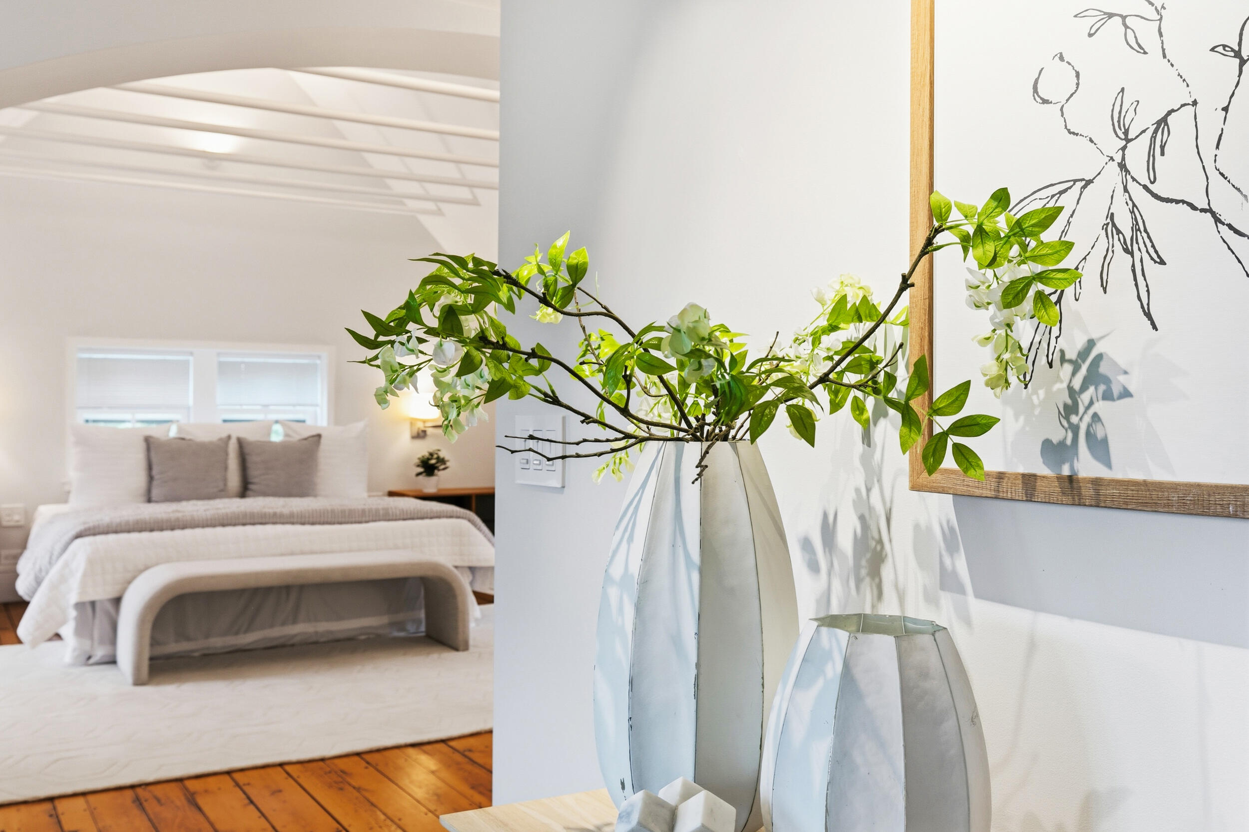 Staged bedroom with a white bed, exposed wood floors, and a vase of flowering branches.