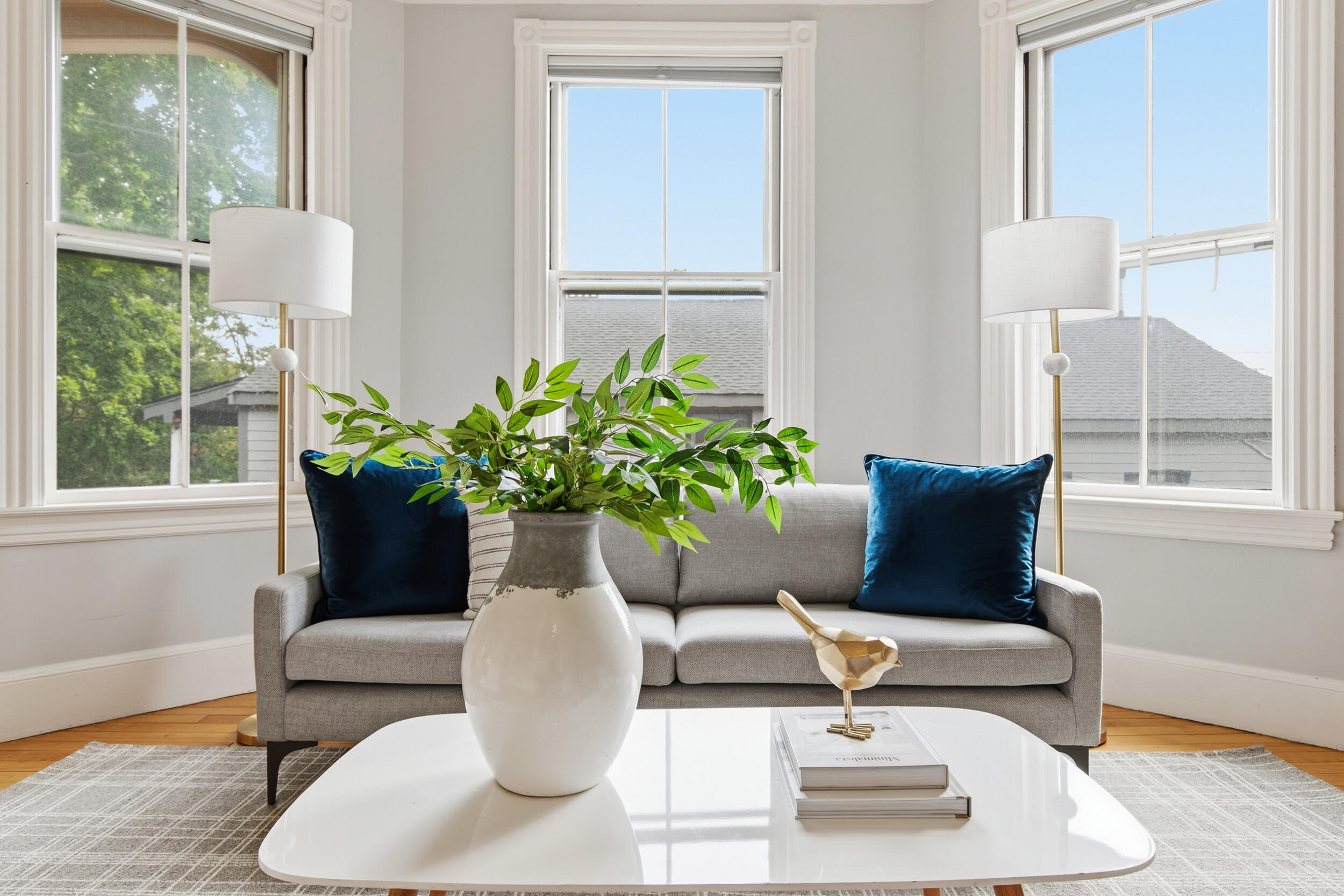 Bright living room with a gray sofa, blue cushions, and tall bay windows.