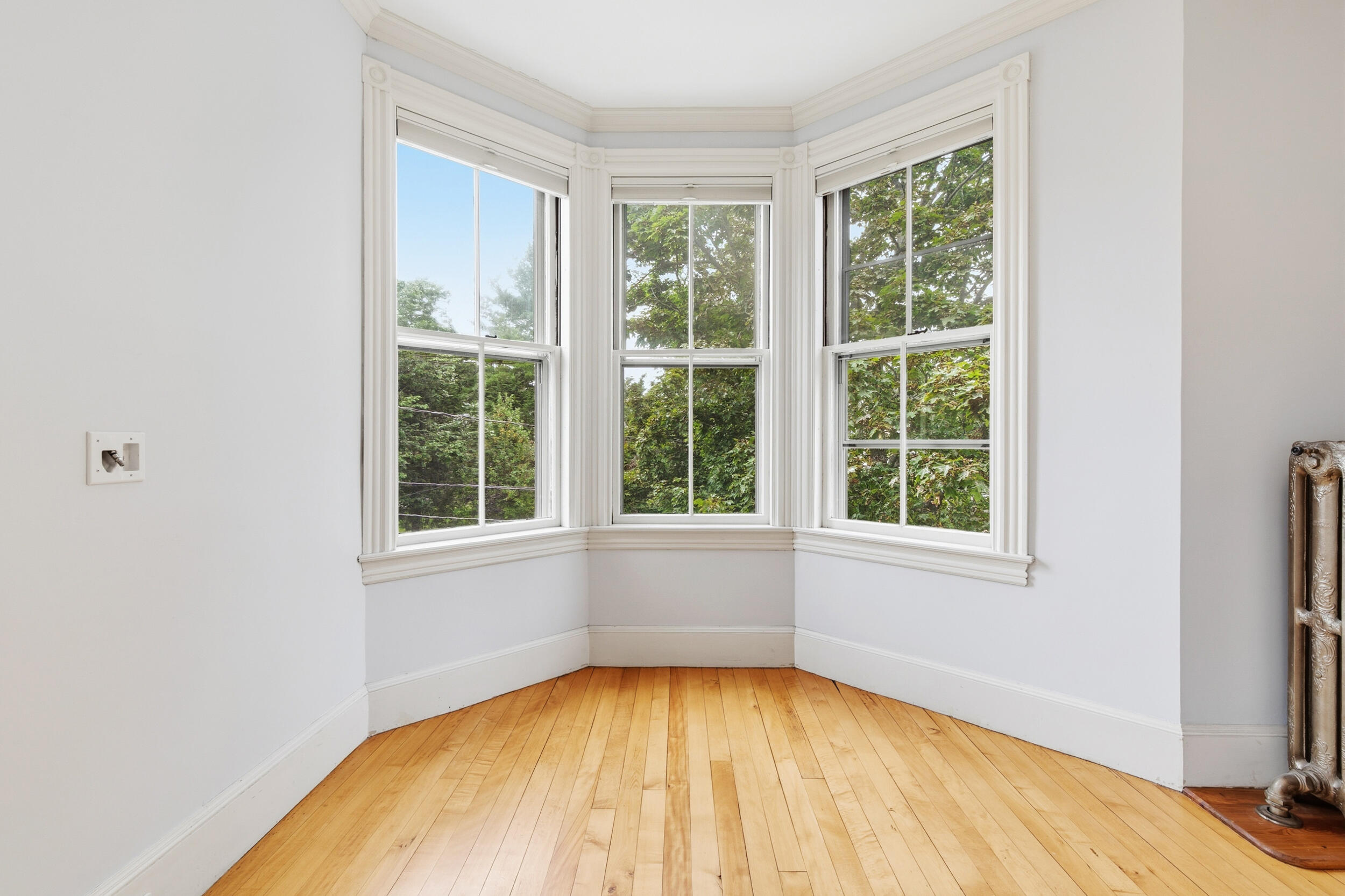 Bright room with a bay of tall windows, hardwood floors, and an ornate cast-iron radiator.