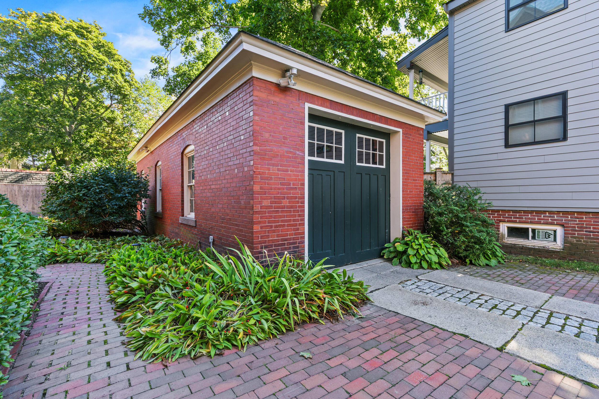 Red brick garage with dark green doors, surrounded by leafy plantings and brick paths.