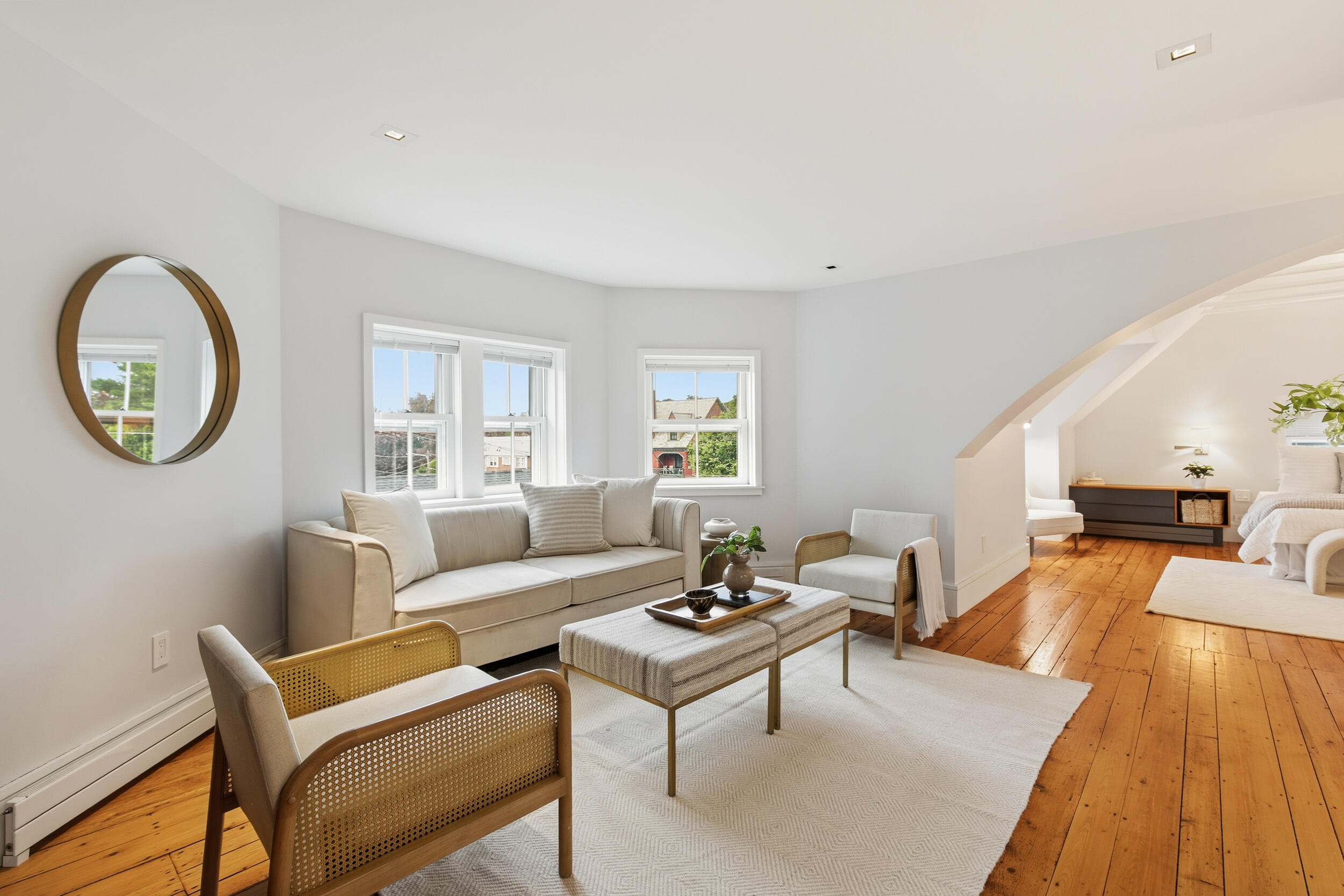 Bright living room with white furnishings, warm wood floors, and an adjoining bedroom alcove.