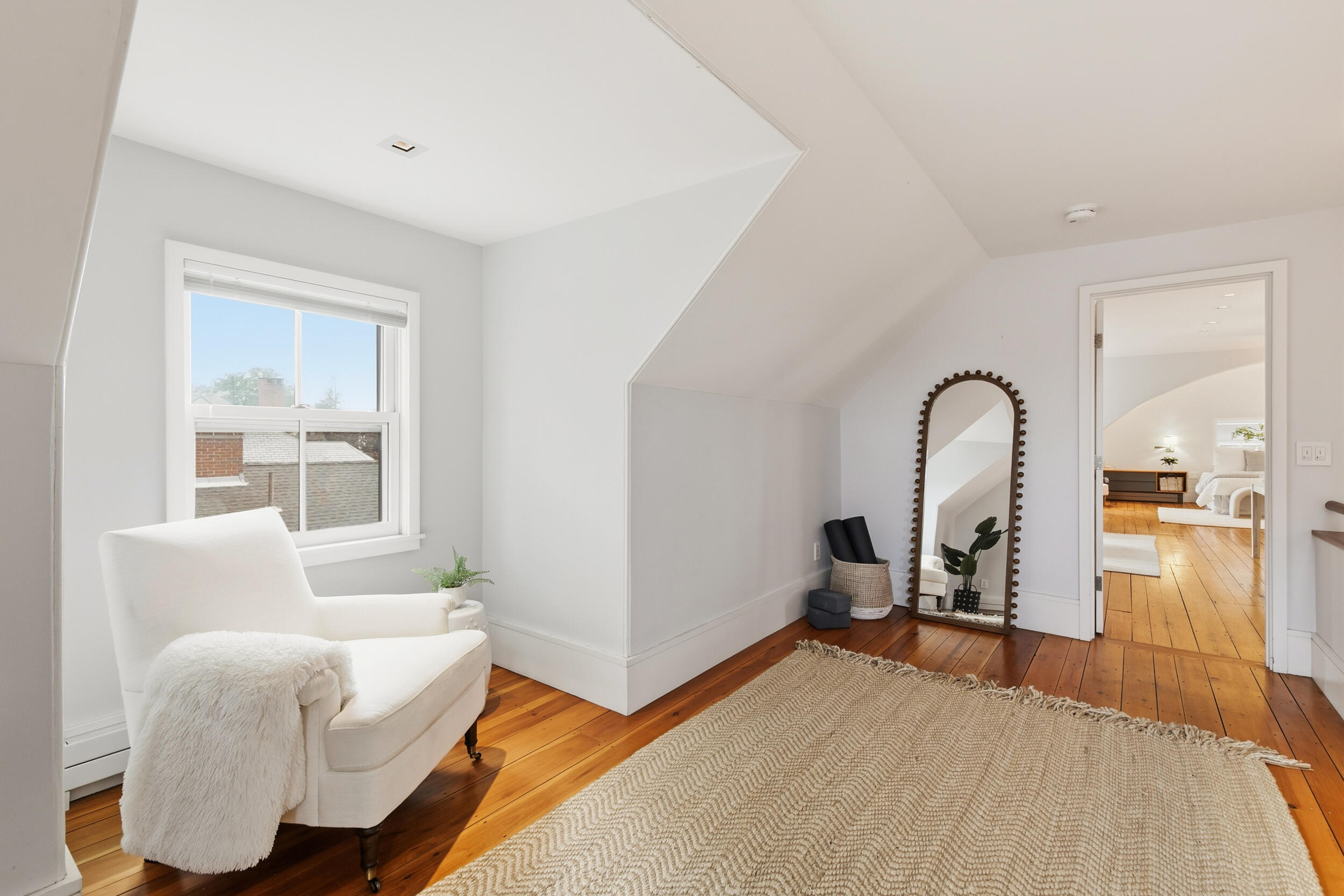 Bright sitting area with a white armchair, exposed wood floors, and a doorway to a bedroom.