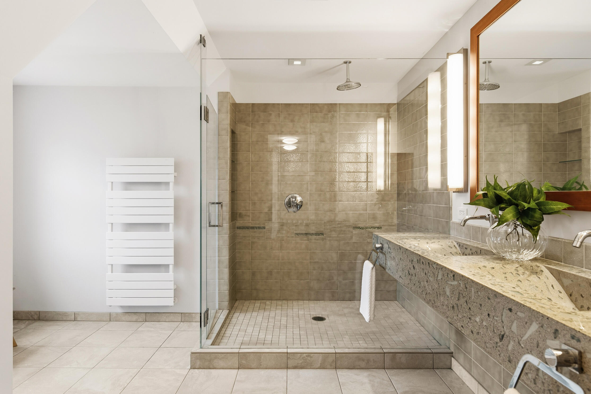 Walk-in tiled shower beside a long stone vanity with twin sinks and a wood-framed mirror.