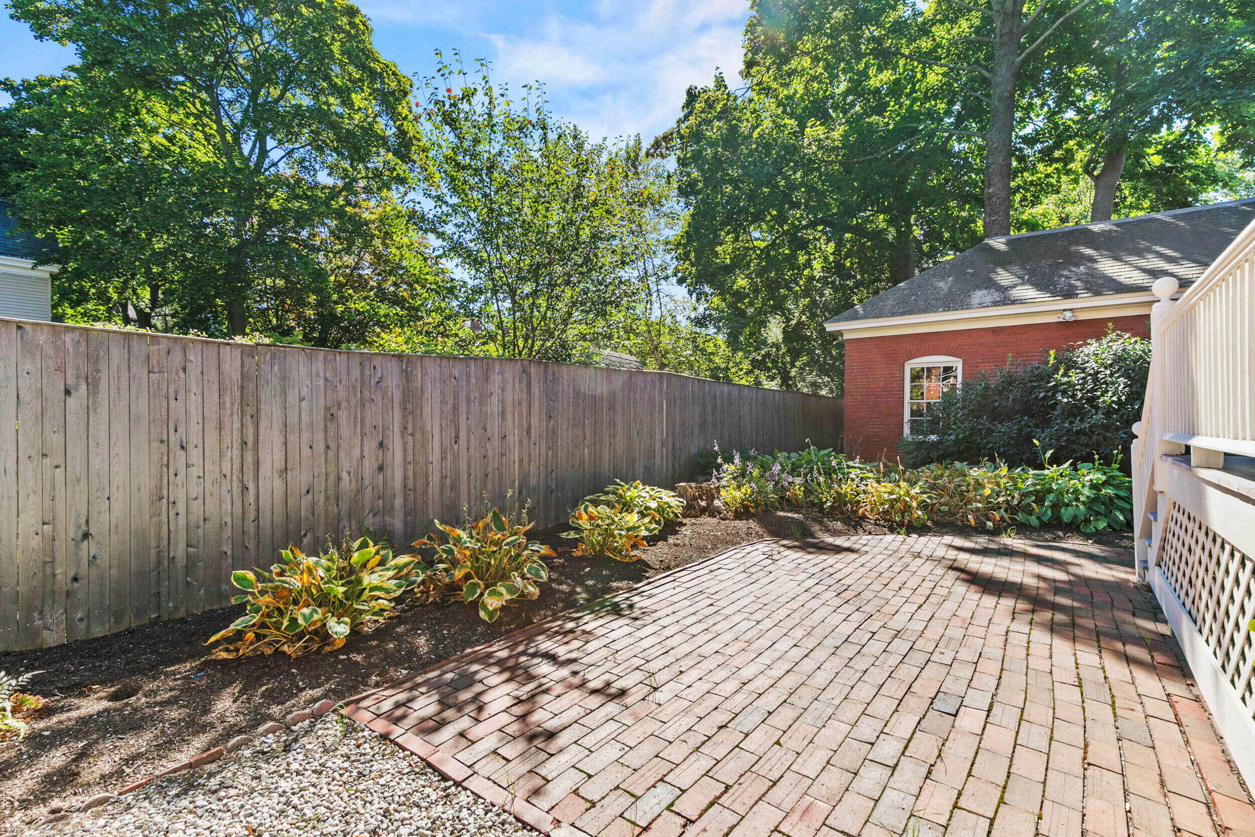 Brick-paved patio bordered by garden beds and a tall wooden fence beneath mature trees.