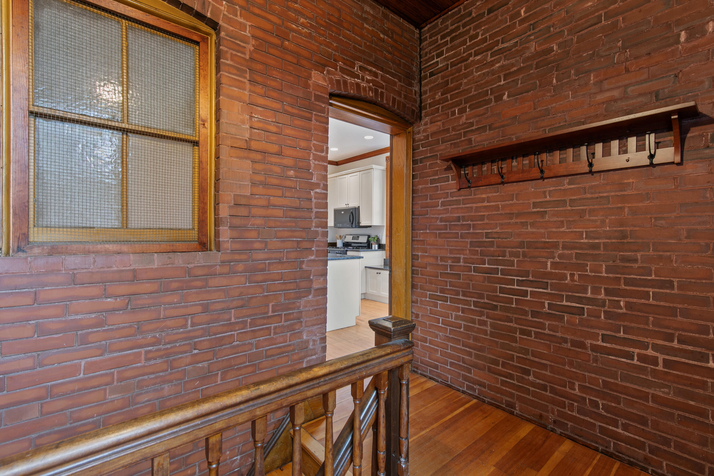 Brick-walled landing with a wooden railing, coat rack, and doorway into a bright kitchen.