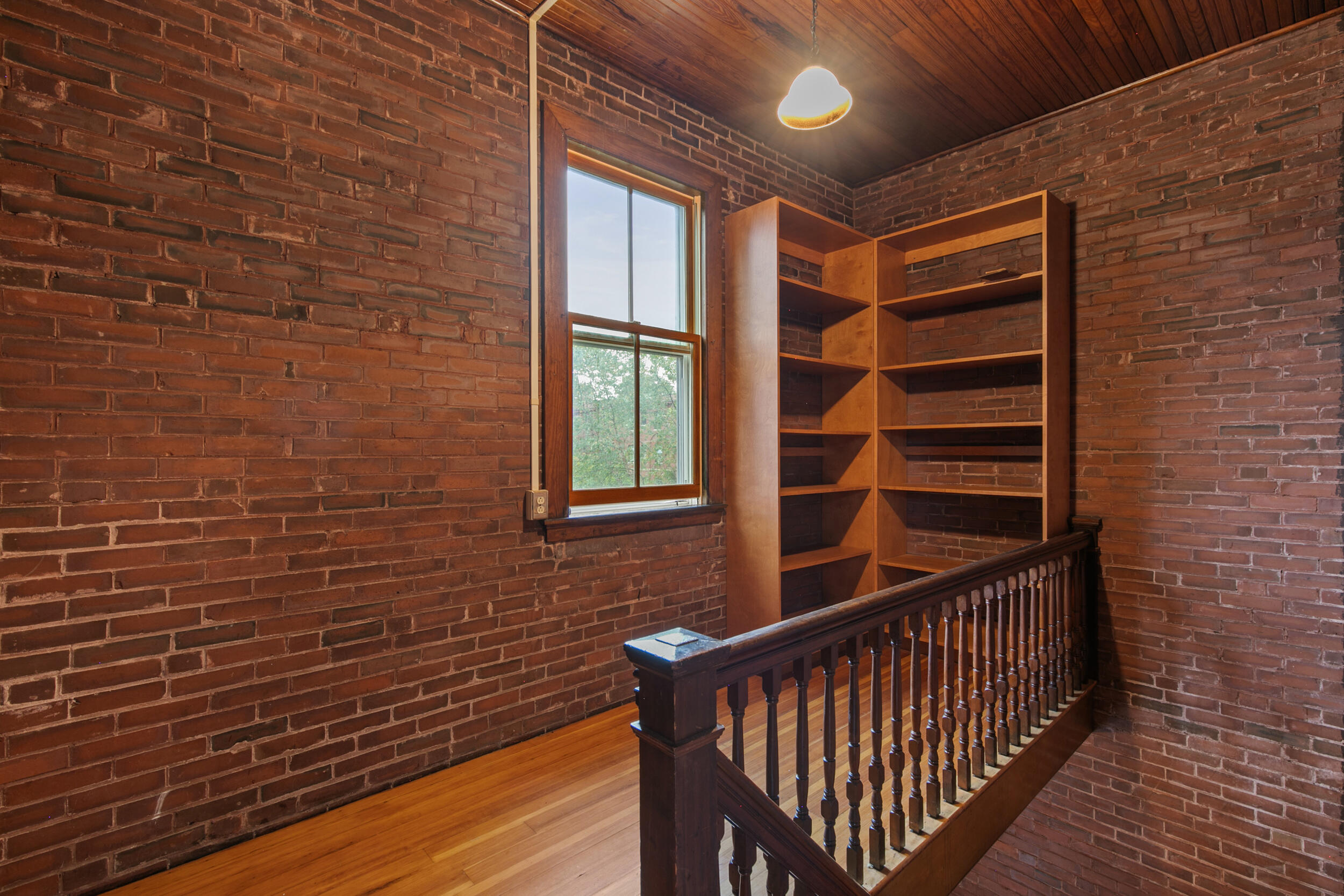 Brick-walled upper landing with built-in wooden shelves, a tall window, and a dark wood staircase railing.