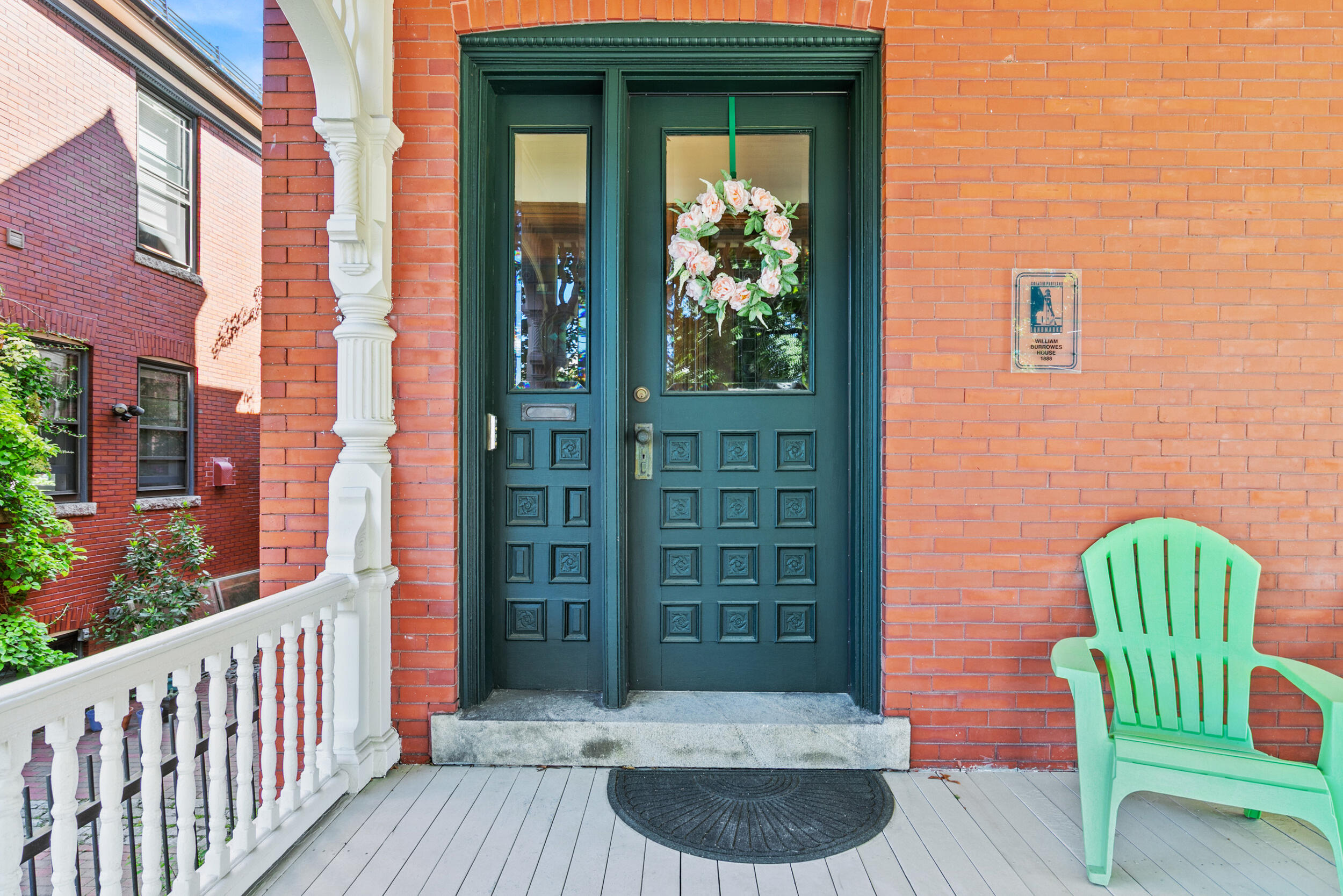 Brick home entrance with a dark green door, floral wreath, white porch railing, and mint-green chair.