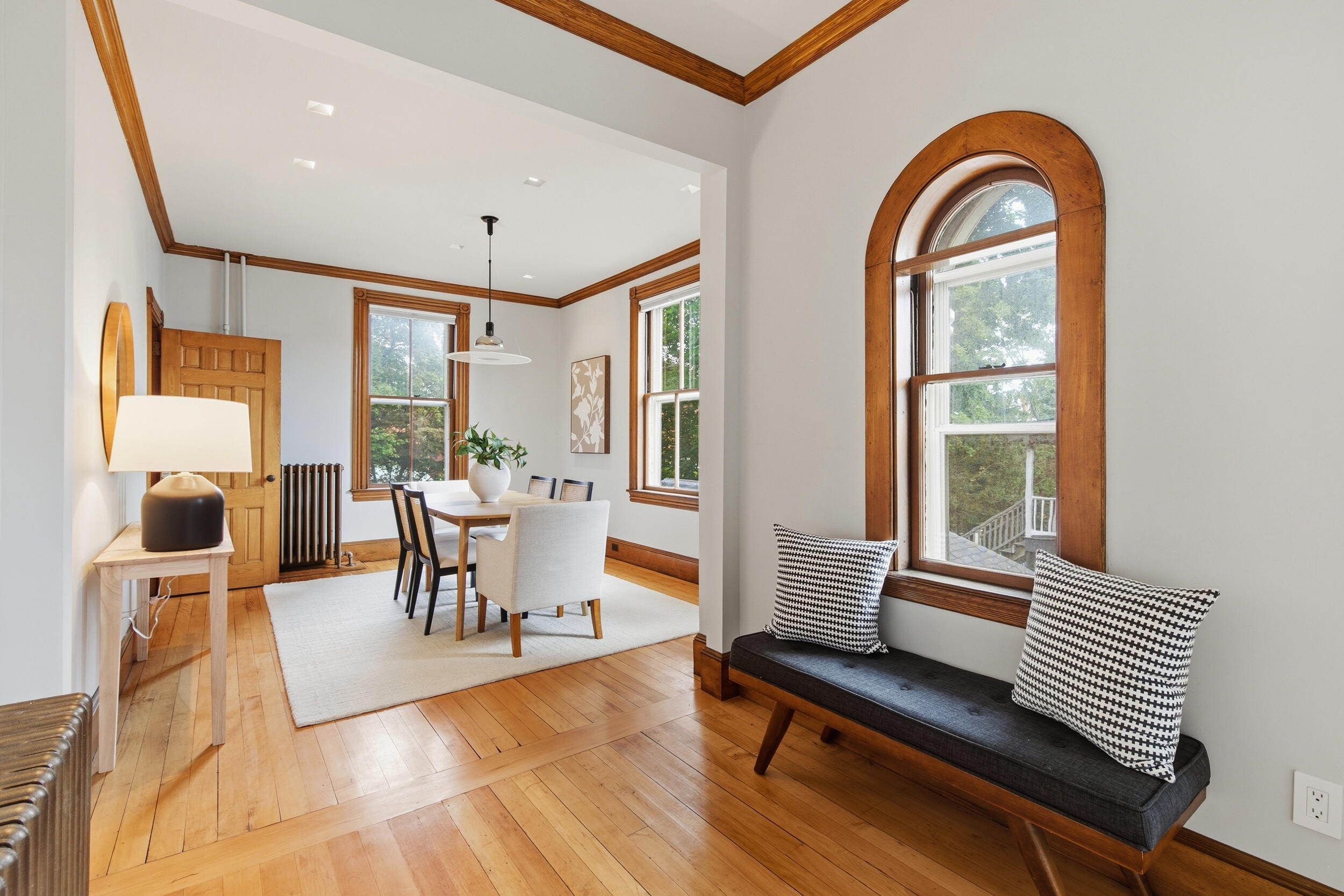 Dining room with hardwood floors, wood-trimmed windows, and a bench beneath an arched window.
