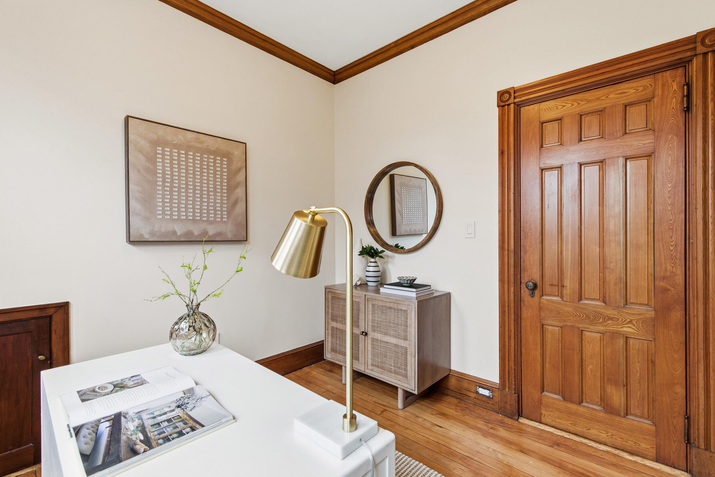 Staged office with a white desk, brass lamp, round mirror, and ornate wooden door.