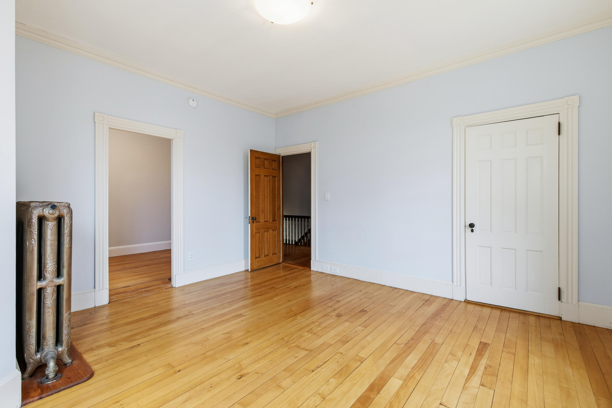 Empty room with pale blue walls, hardwood floors, ornate trim, and an antique radiator.