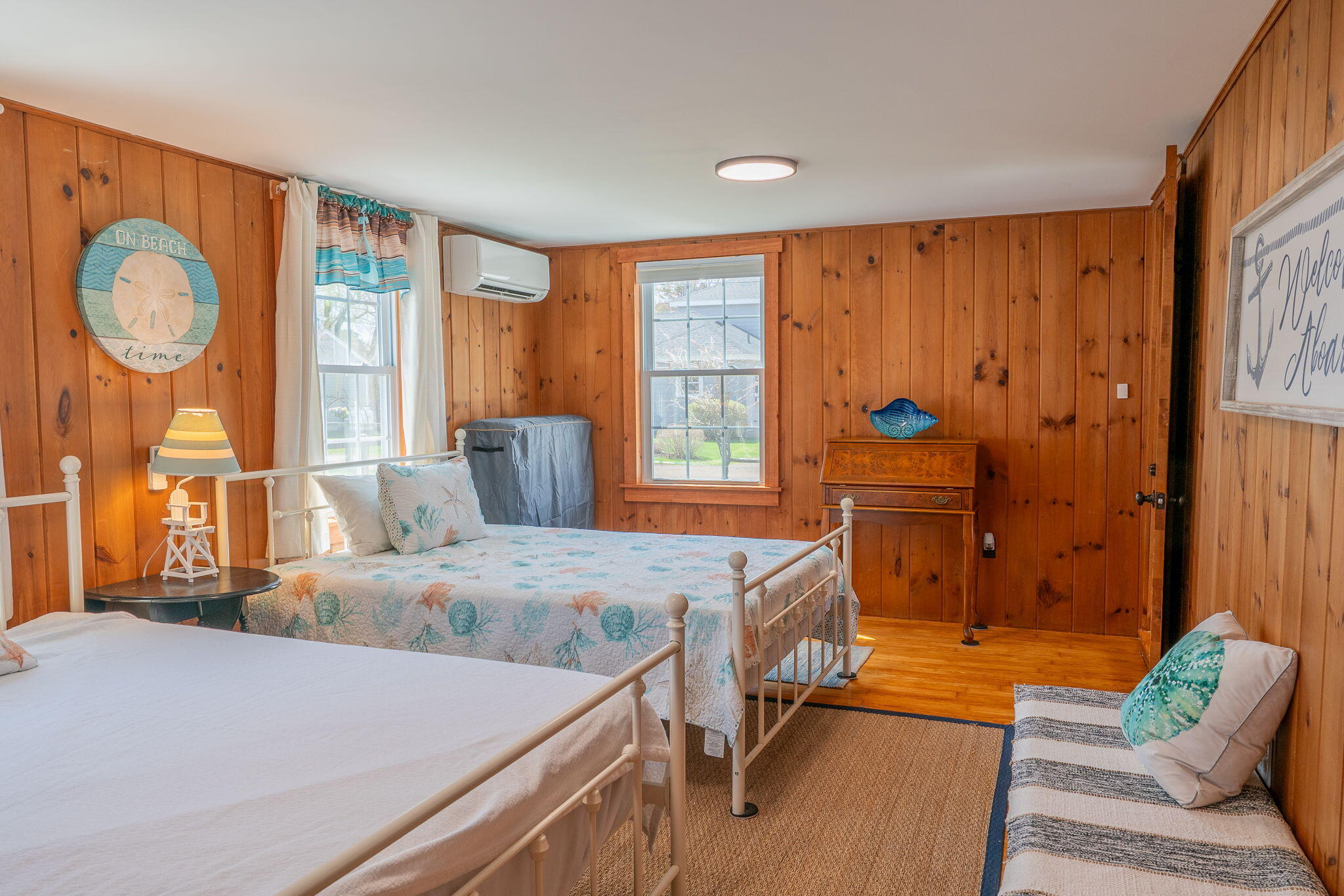 Wood-paneled bedroom with two white metal beds, a coastal quilt, and a striped loveseat.