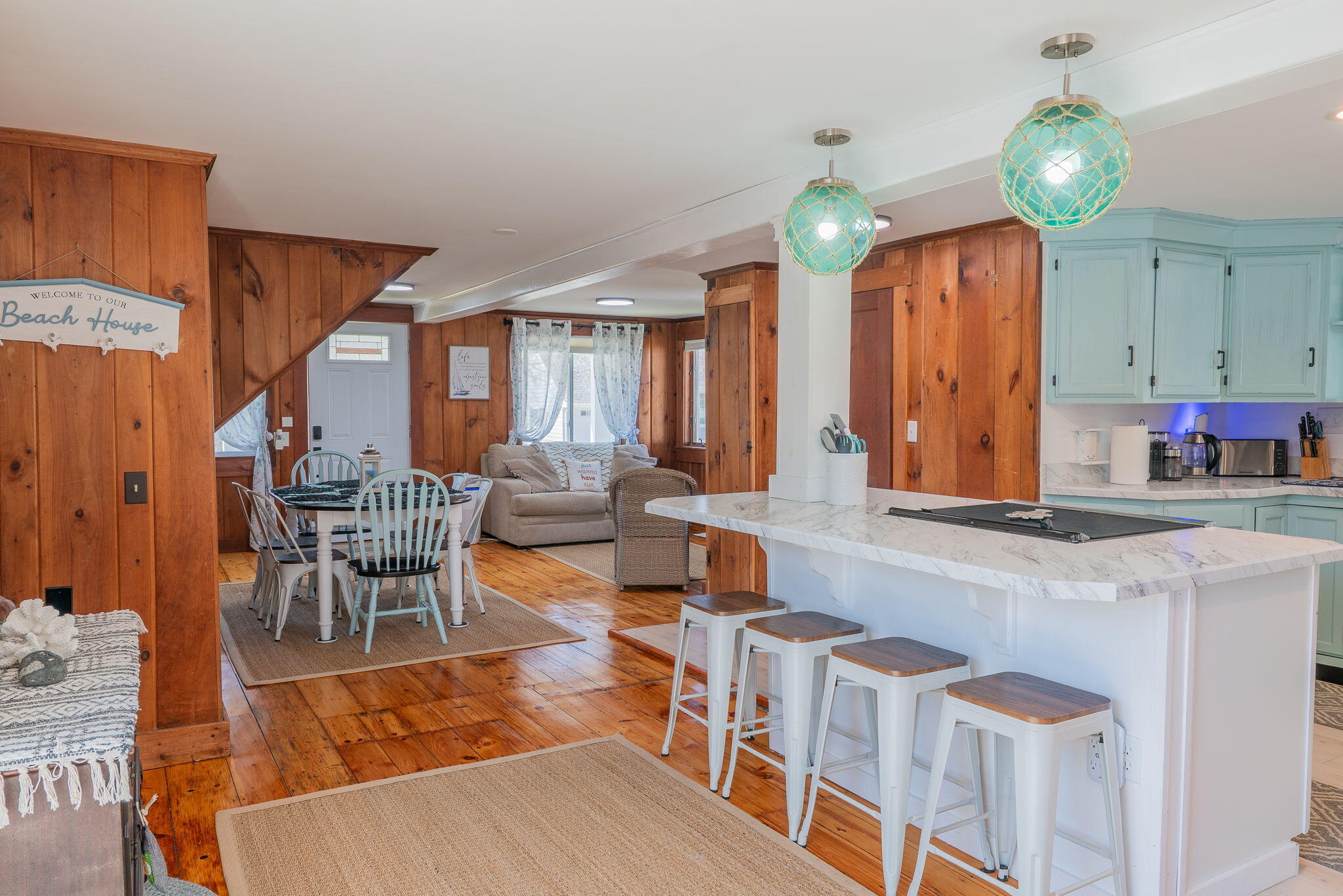Open-plan kitchen and living room with a marble island, turquoise pendant lights, and warm wood walls.