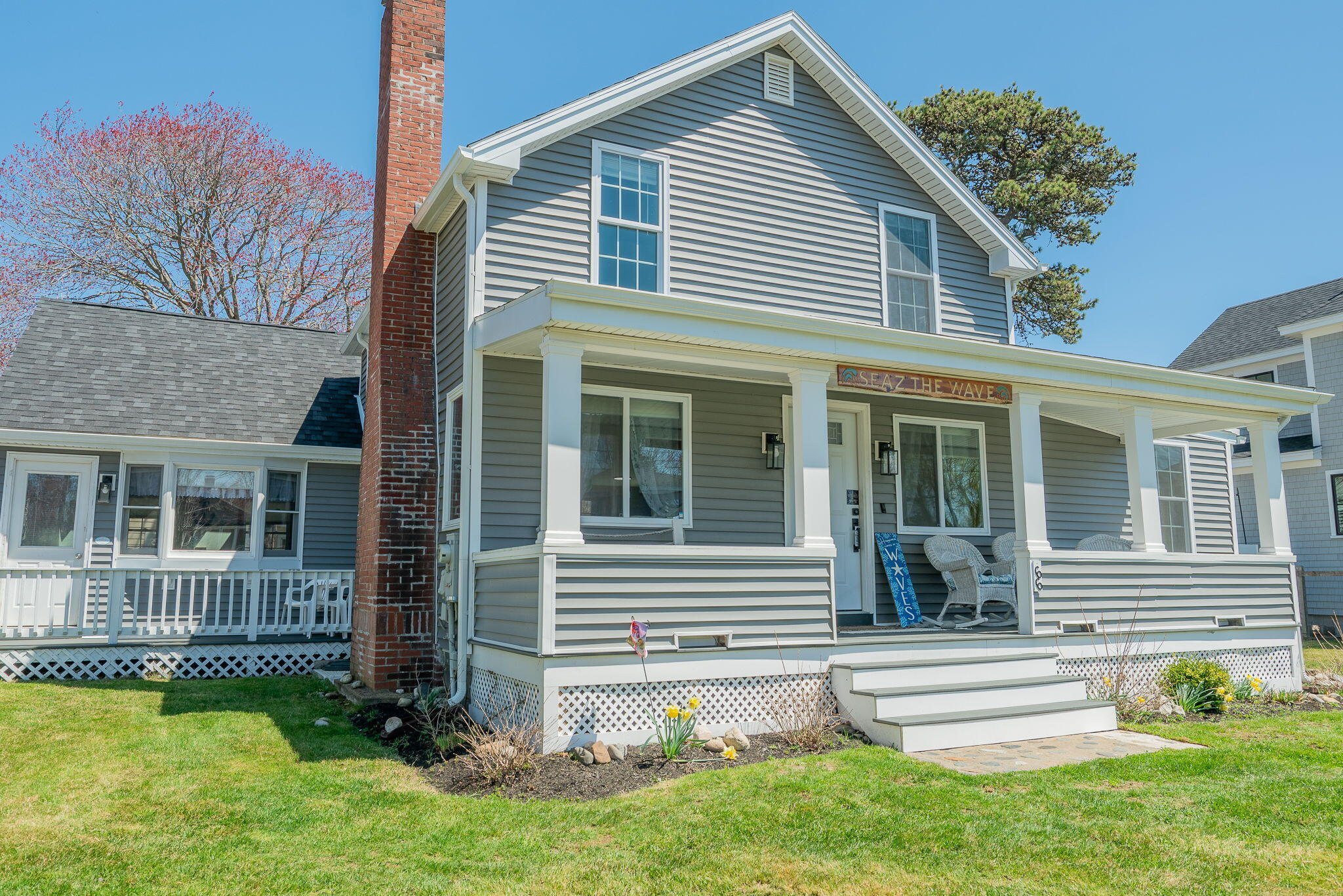 Gray two-story house with a broad covered porch, brick chimney, and spring flowers.