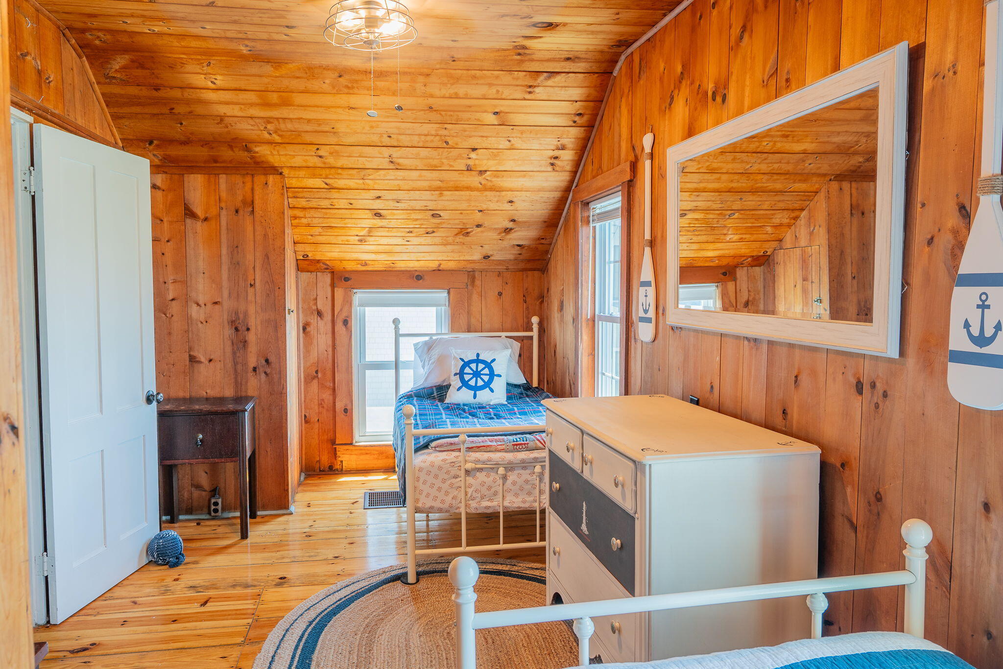 Wood-paneled bedroom with sloped ceiling, nautical bedding, two beds, and a white dresser.