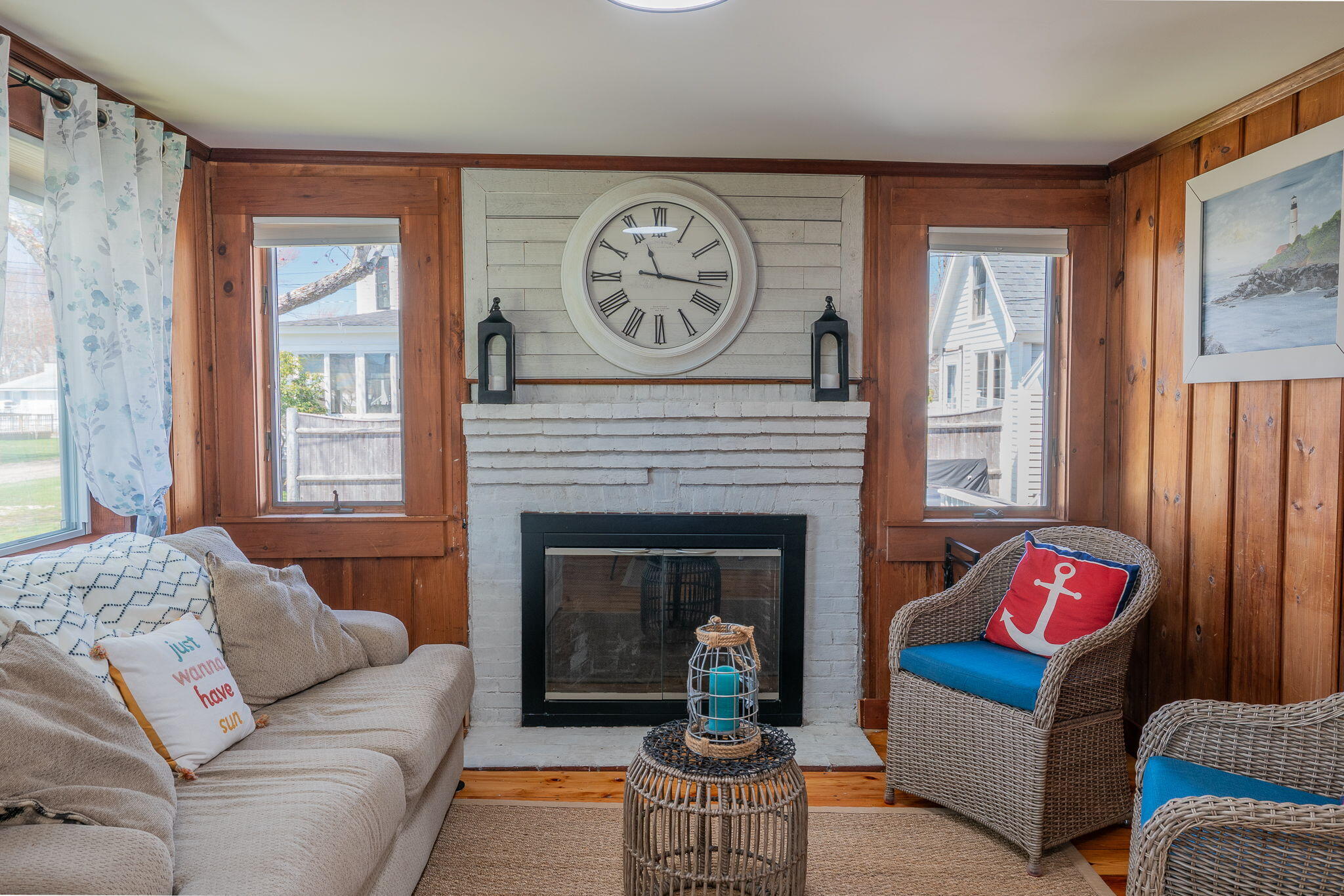 Living room with a white brick fireplace, wood-paneled walls, and wicker chairs with blue cushions.