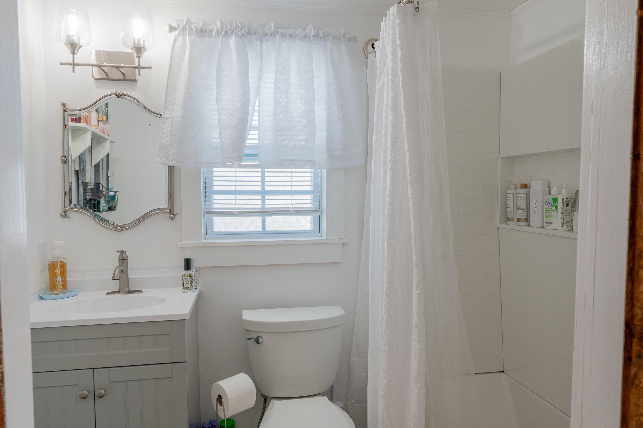 Bathroom with a gray vanity, window, toilet, and white-tiled shower.