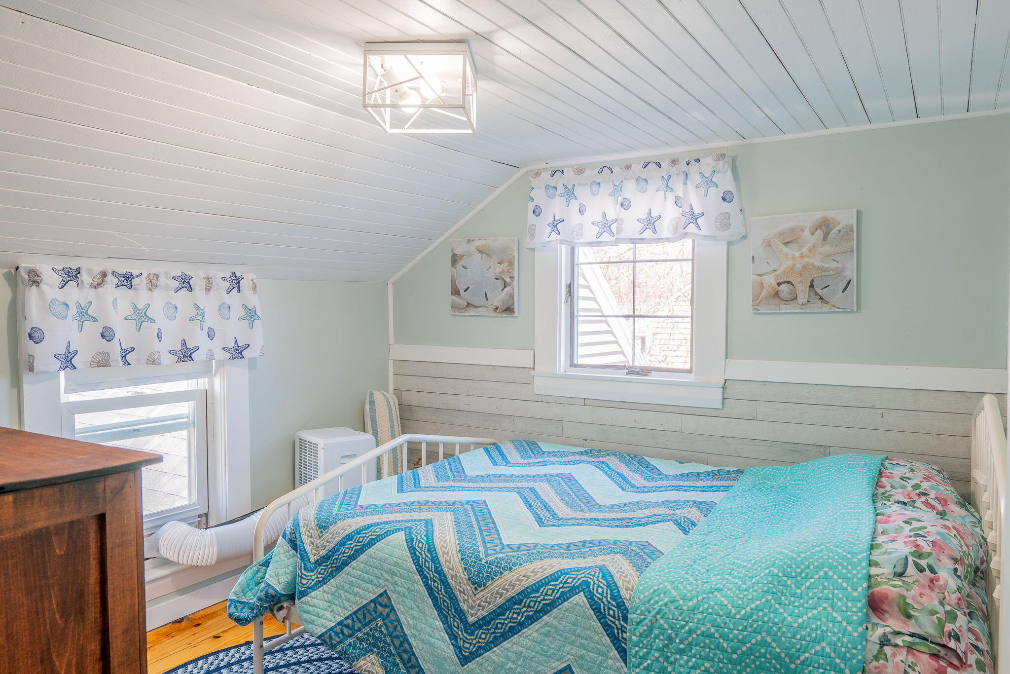 Bedroom with a sloped plank ceiling, blue quilts, and starfish-patterned curtains.