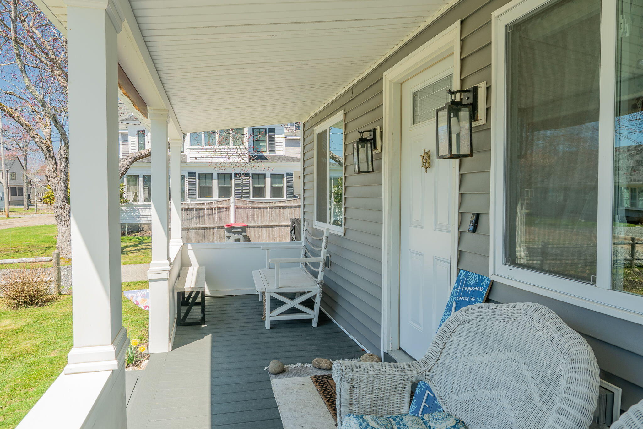 Covered porch with white wicker seating, gray siding, and a row of columns beside the lawn.