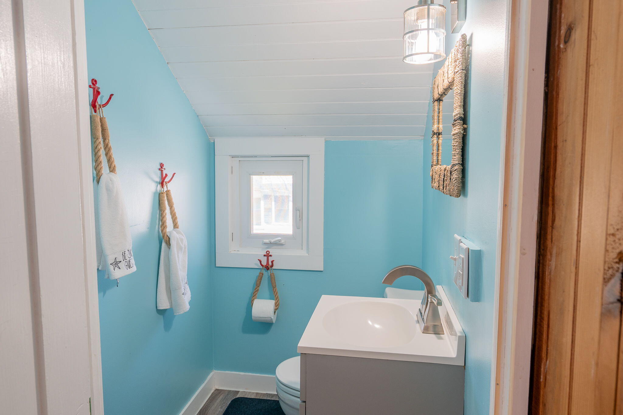 Turquoise bathroom with a white vanity and toilet, nautical rope hooks, and a small window.