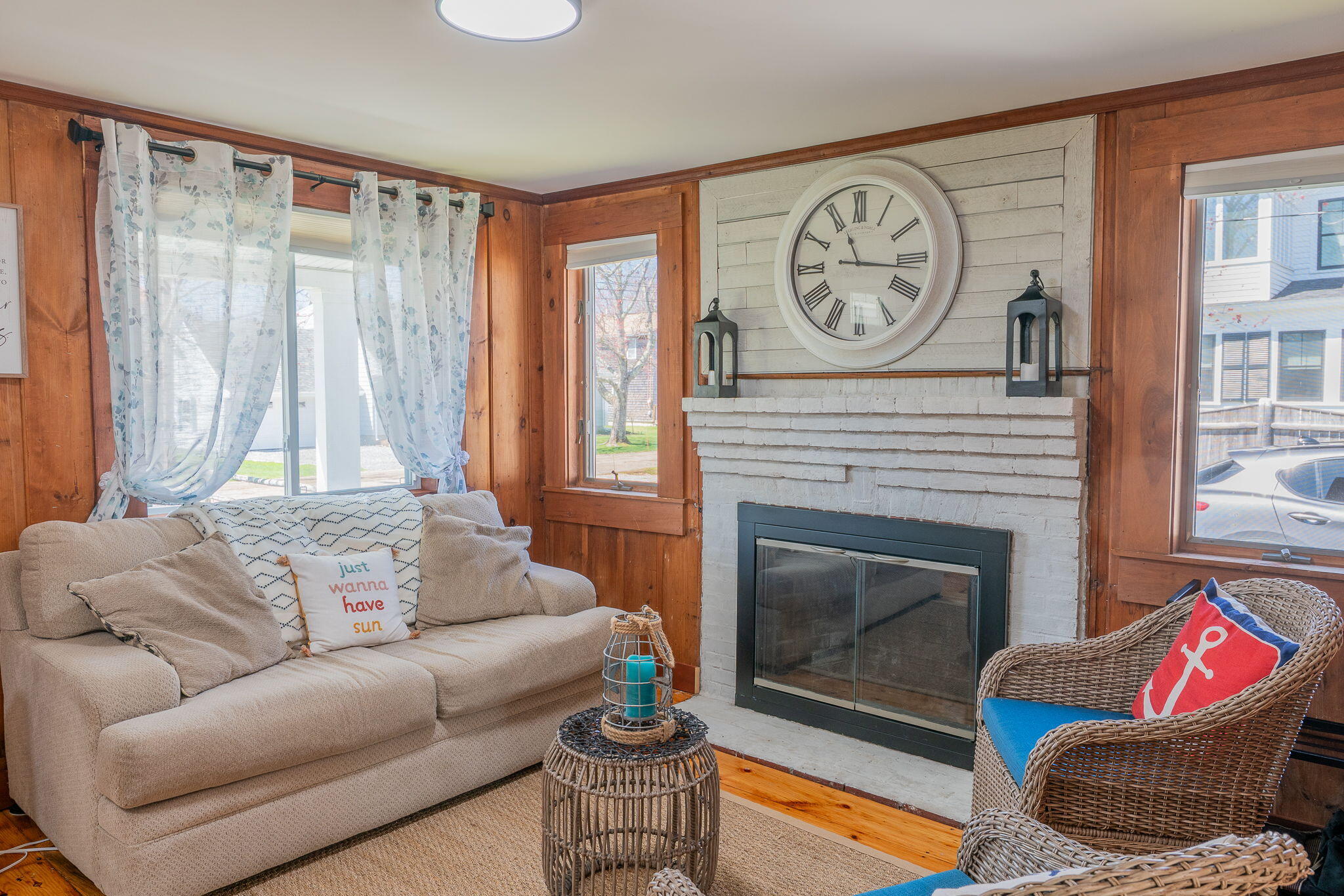 Living room with a white brick fireplace, oversized clock, beige sectional, and wicker chair.