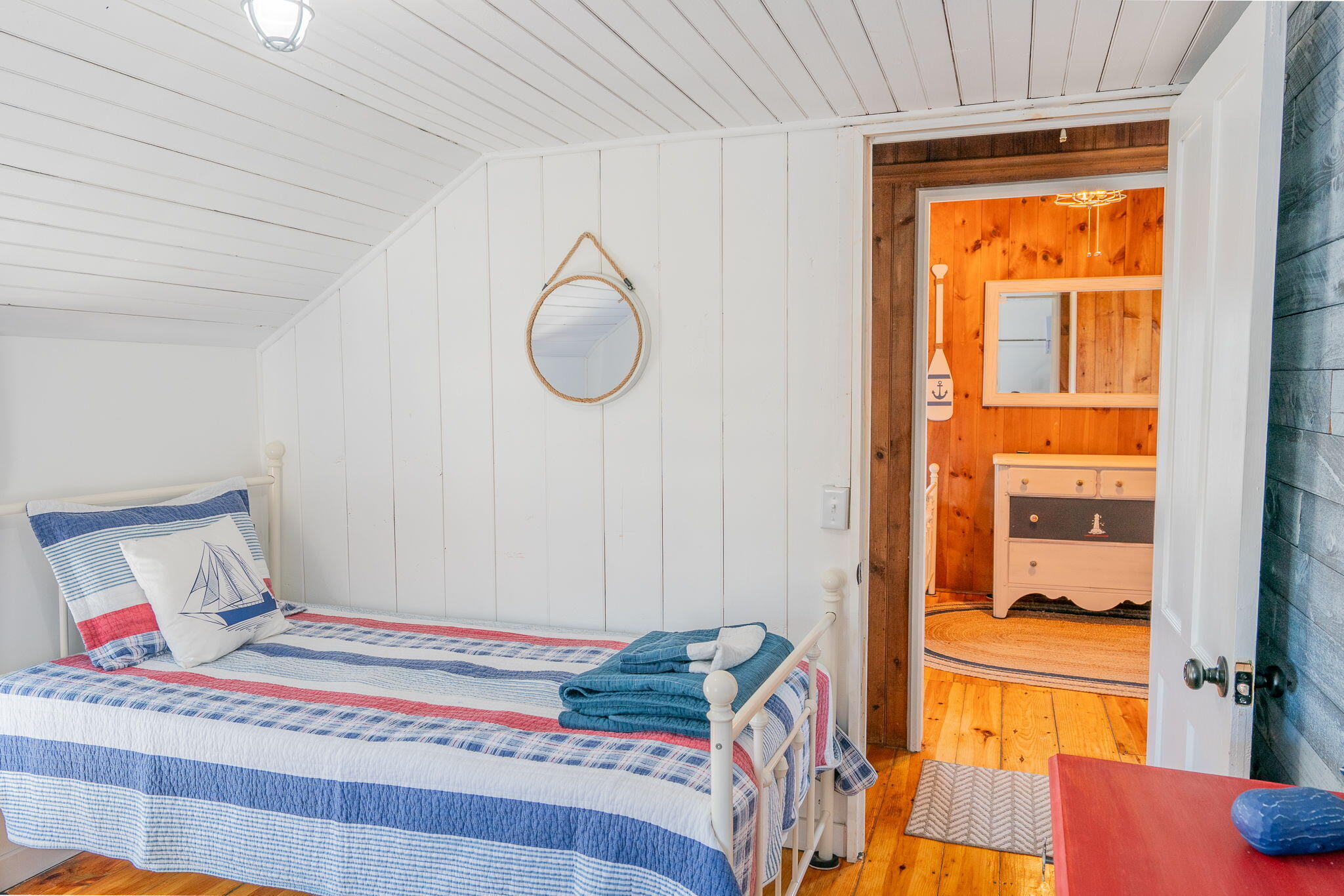Bedroom with striped nautical bedding, white beadboard walls, and an open doorway to a wood-paneled room.