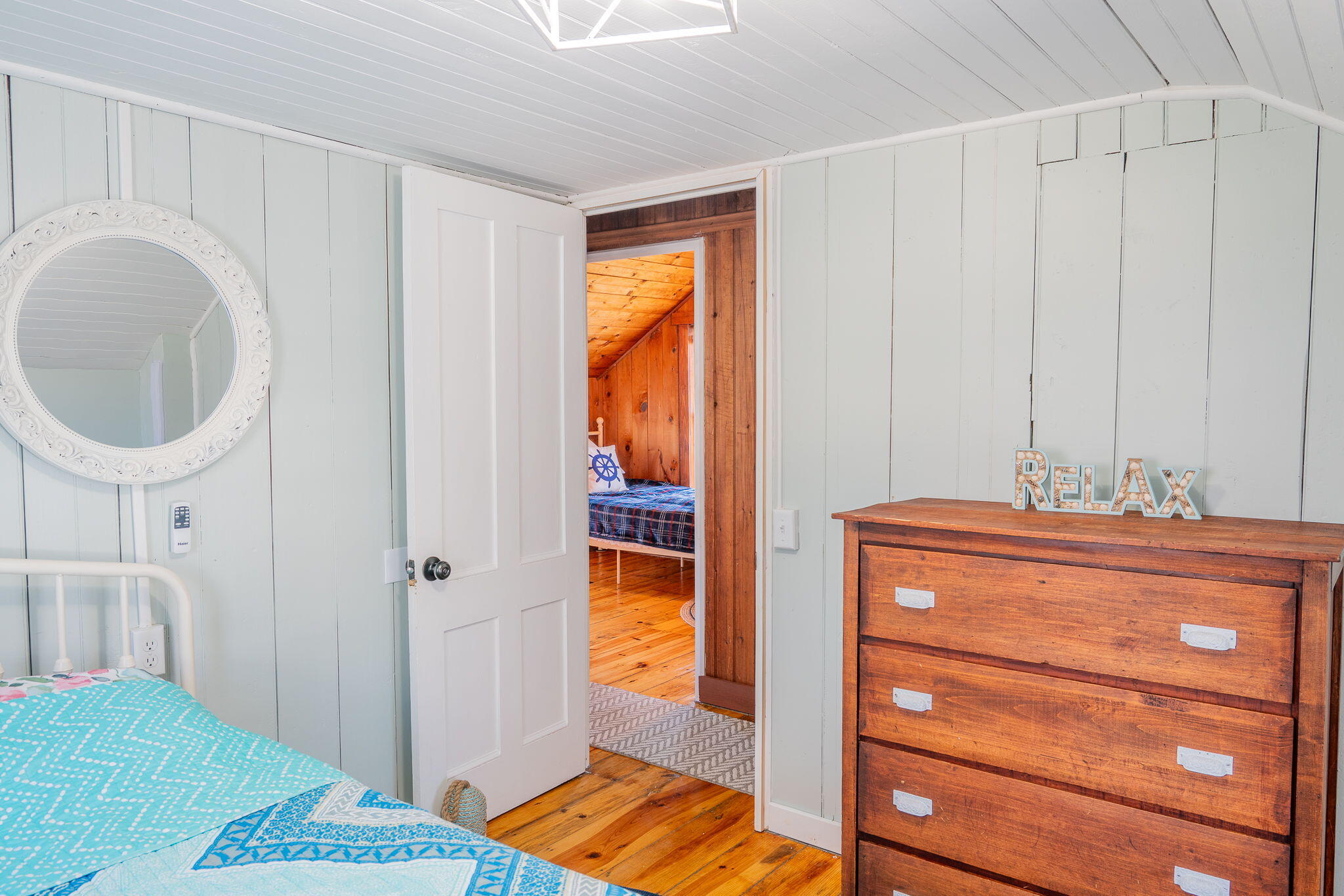 Bedroom with a turquoise-covered bed and dresser, looking through an open doorway into a wood-paneled bedroom.