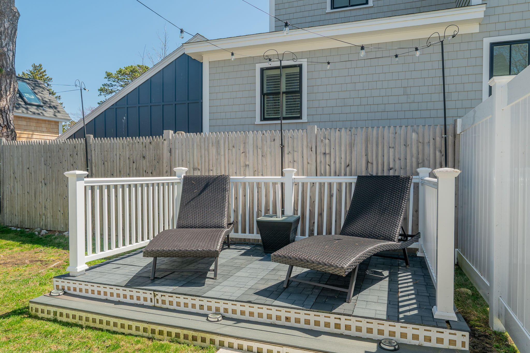 Outdoor deck with two woven chaise lounges, gray pavers, white railings, and a tall privacy fence.