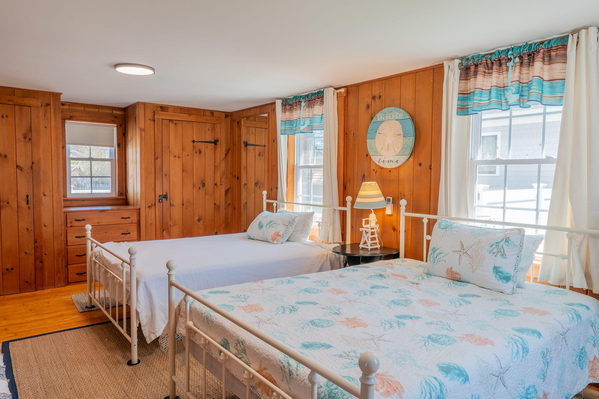 Bedroom with two seashell-patterned beds, pine-paneled walls, and built-in drawers beneath a window.