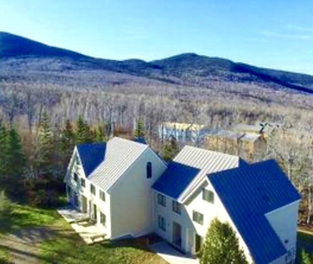 Aerial view of a pale-sided home with blue metal roofs and mountain ridges beyond.